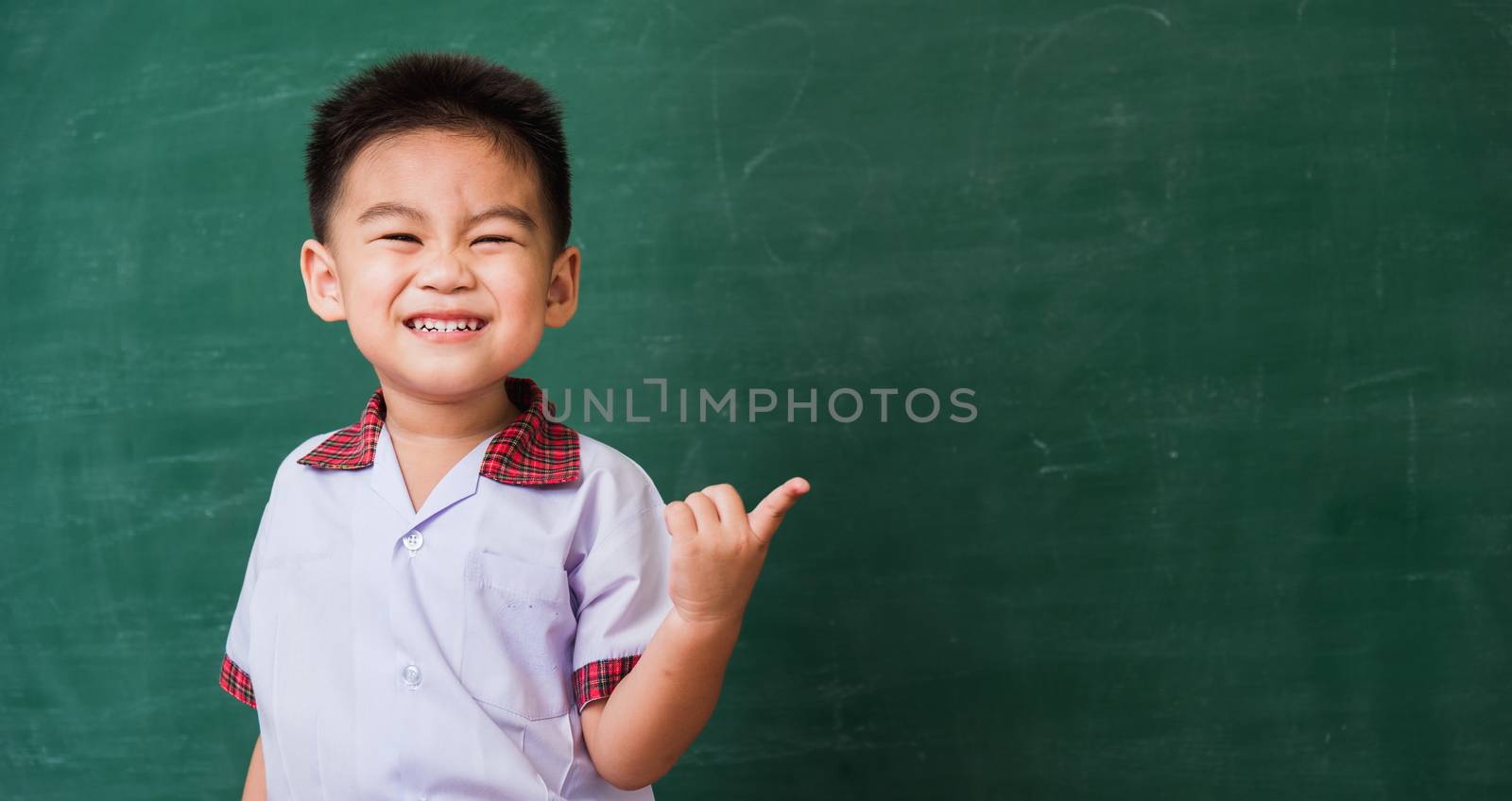 Back to School. Happy Asian funny cute little child boy from kindergarten in student uniform smiling point finger to side away space on green school blackboard, First time to school education concept