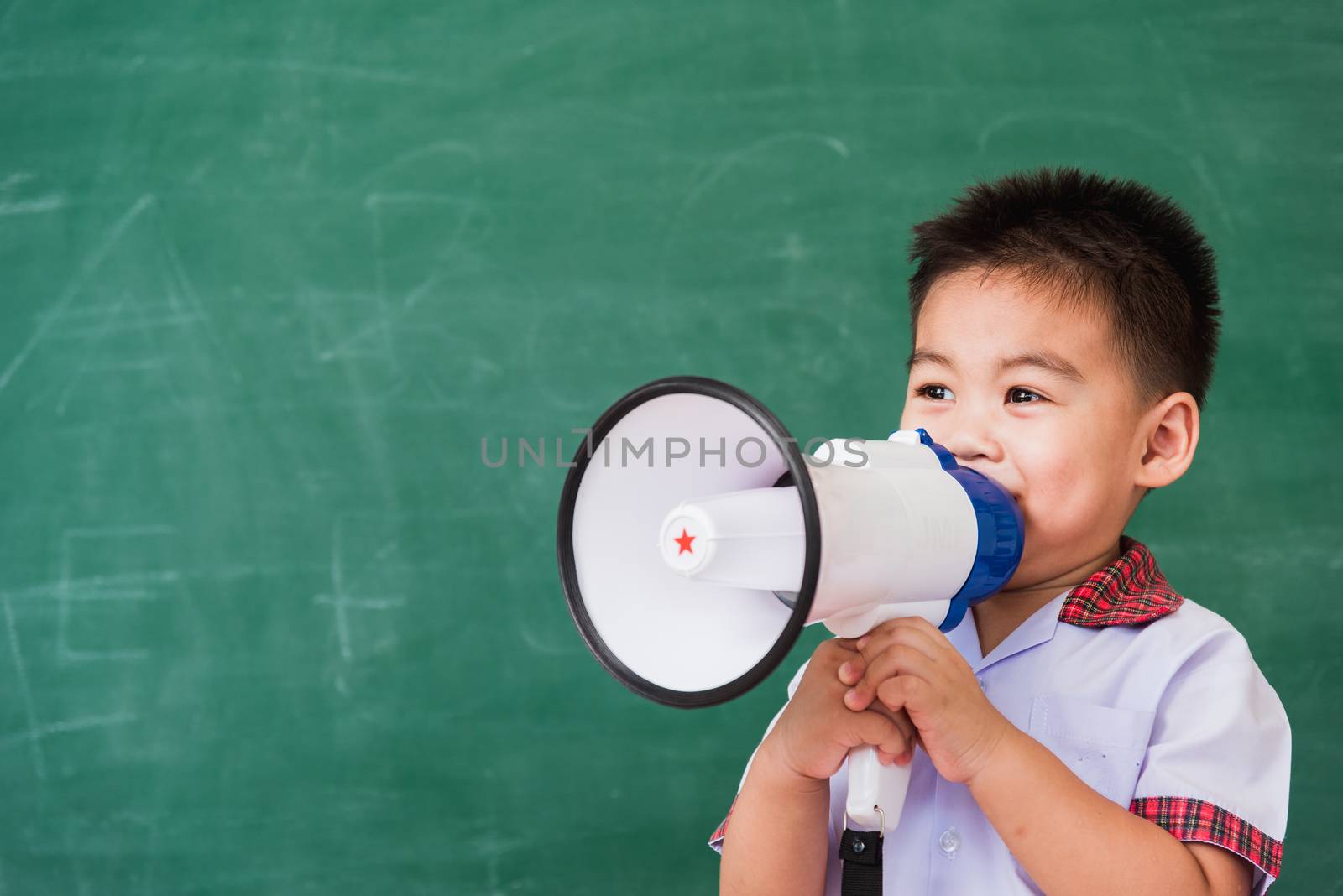 Back to School. Asian funny cute little child boy kindergarten preschool in student uniform speaking through megaphone against on green school blackboard, First time to school education concept