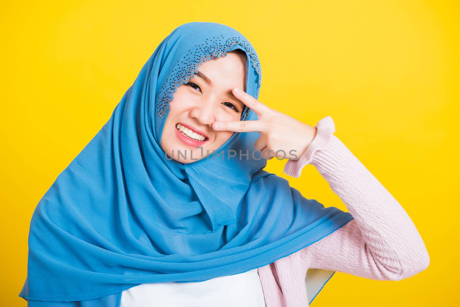 Asian Muslim Arab, Portrait of happy beautiful young woman Islam religious wear veil hijab smile she showing gesture hand two finger V-sign symbol near eyes isolated yellow background