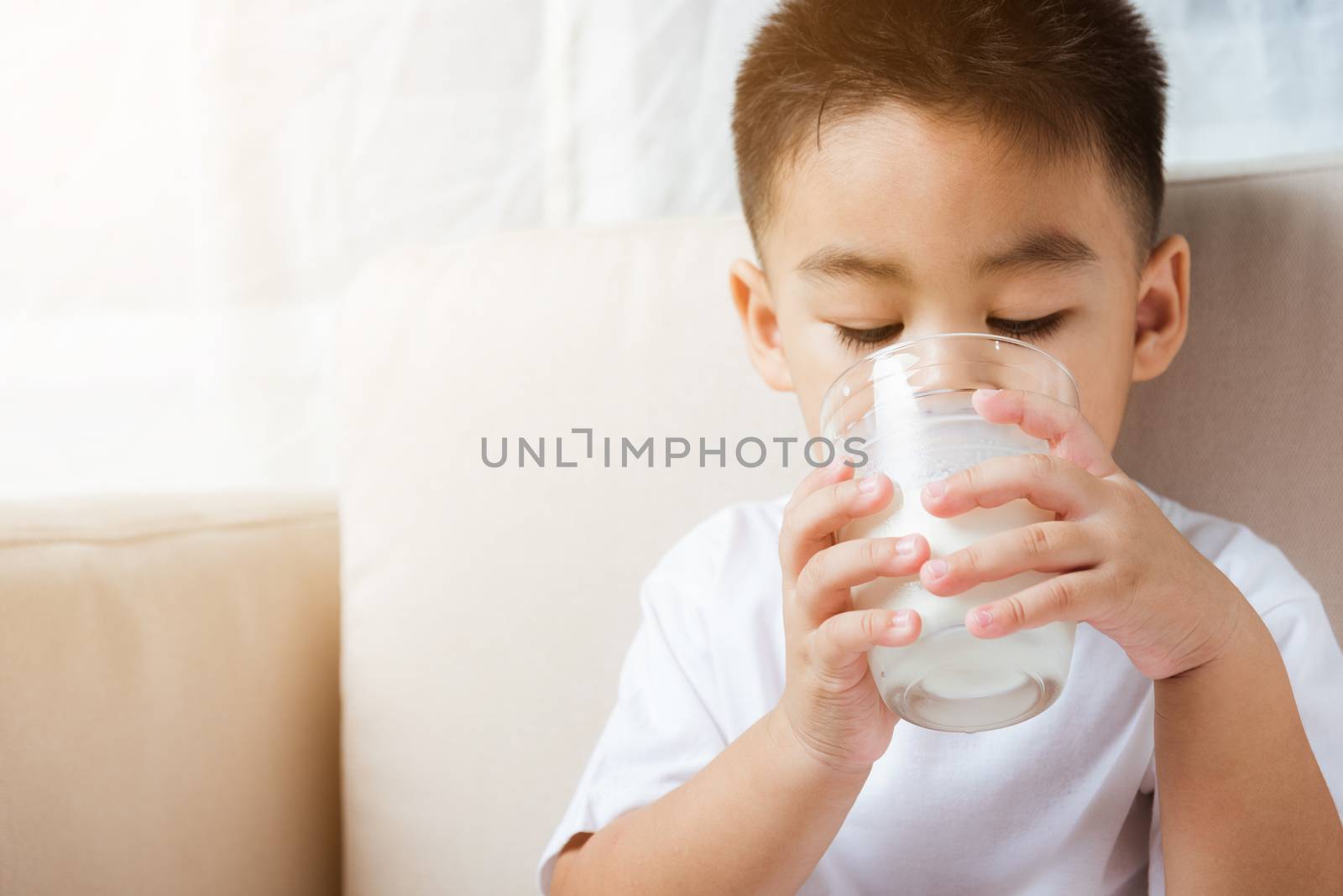 Close up of happy Asian little cute child boy hand holding milk glass he drinking white milk during sitting on the sofa at home after lunch. Daily life health care Medicine food