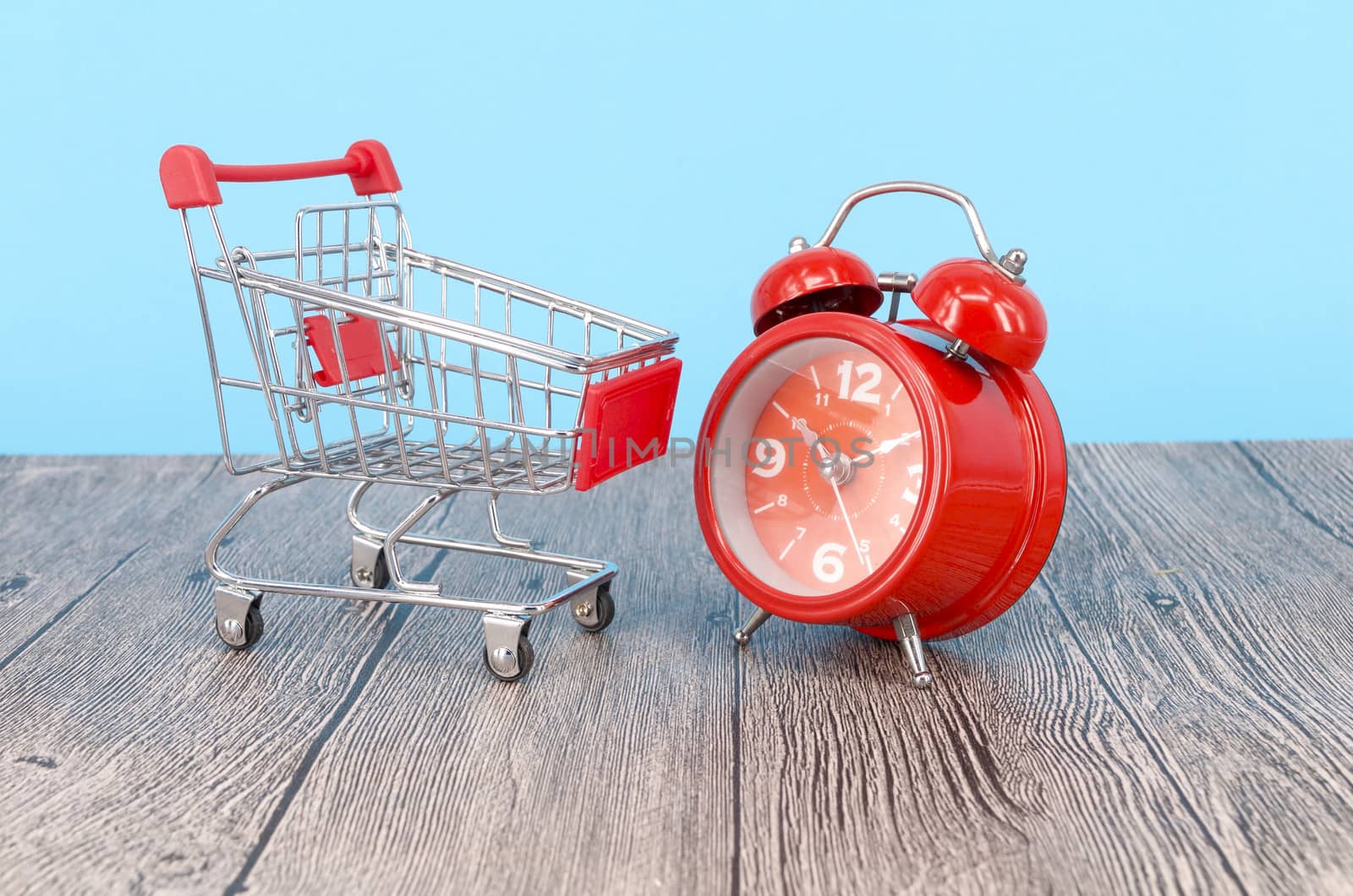 Shopping cart and classic alarm clock on wooden surface. Sale time buy mall market shop consumer concept. Selective focus.