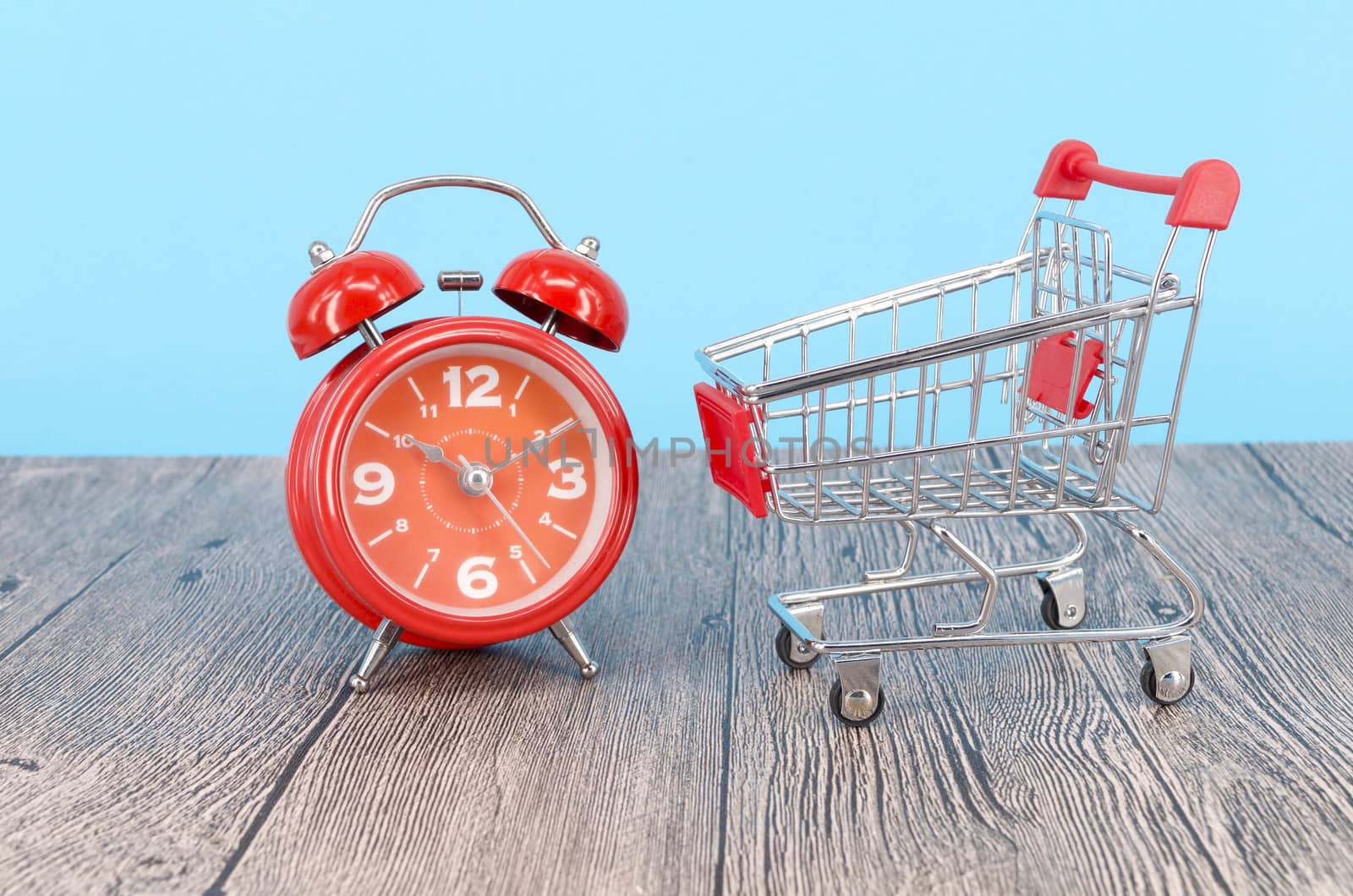 Shopping cart and classic alarm clock on wooden surface. Sale time buy mall market shop consumer concept. Selective focus.