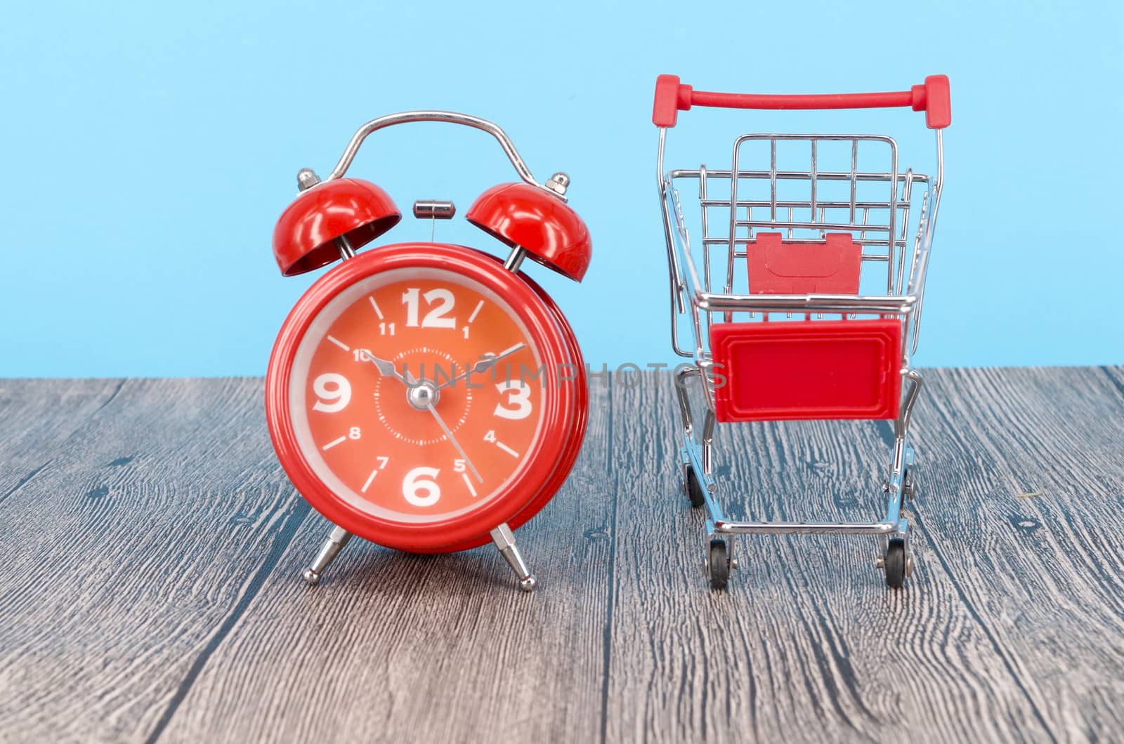 Shopping cart and classic alarm clock on wooden surface. Sale time buy mall market shop consumer concept. Selective focus.