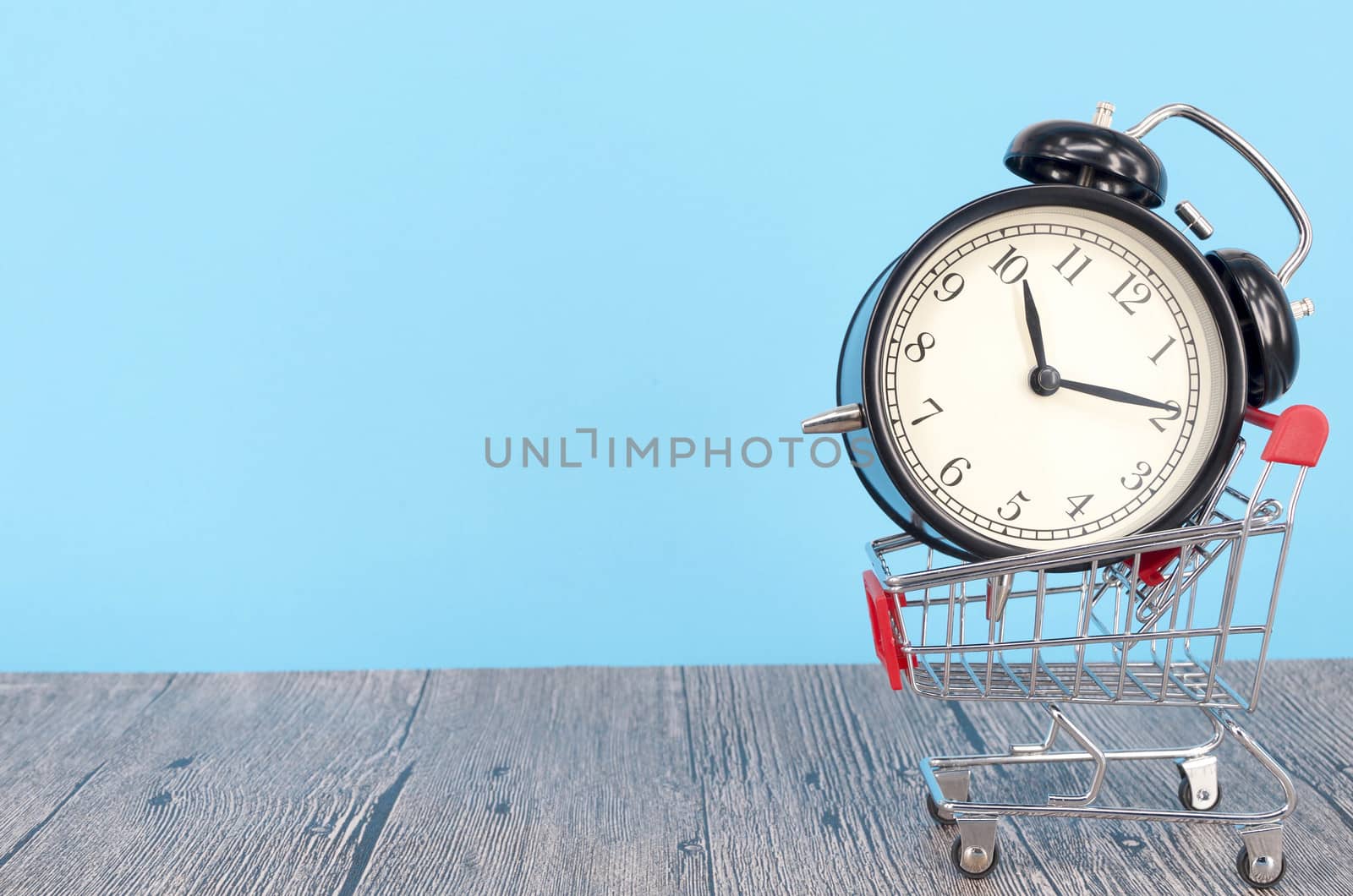 Shopping cart and classic alarm clock on wooden surface. Sale time buy mall market shop consumer concept. Selective focus.