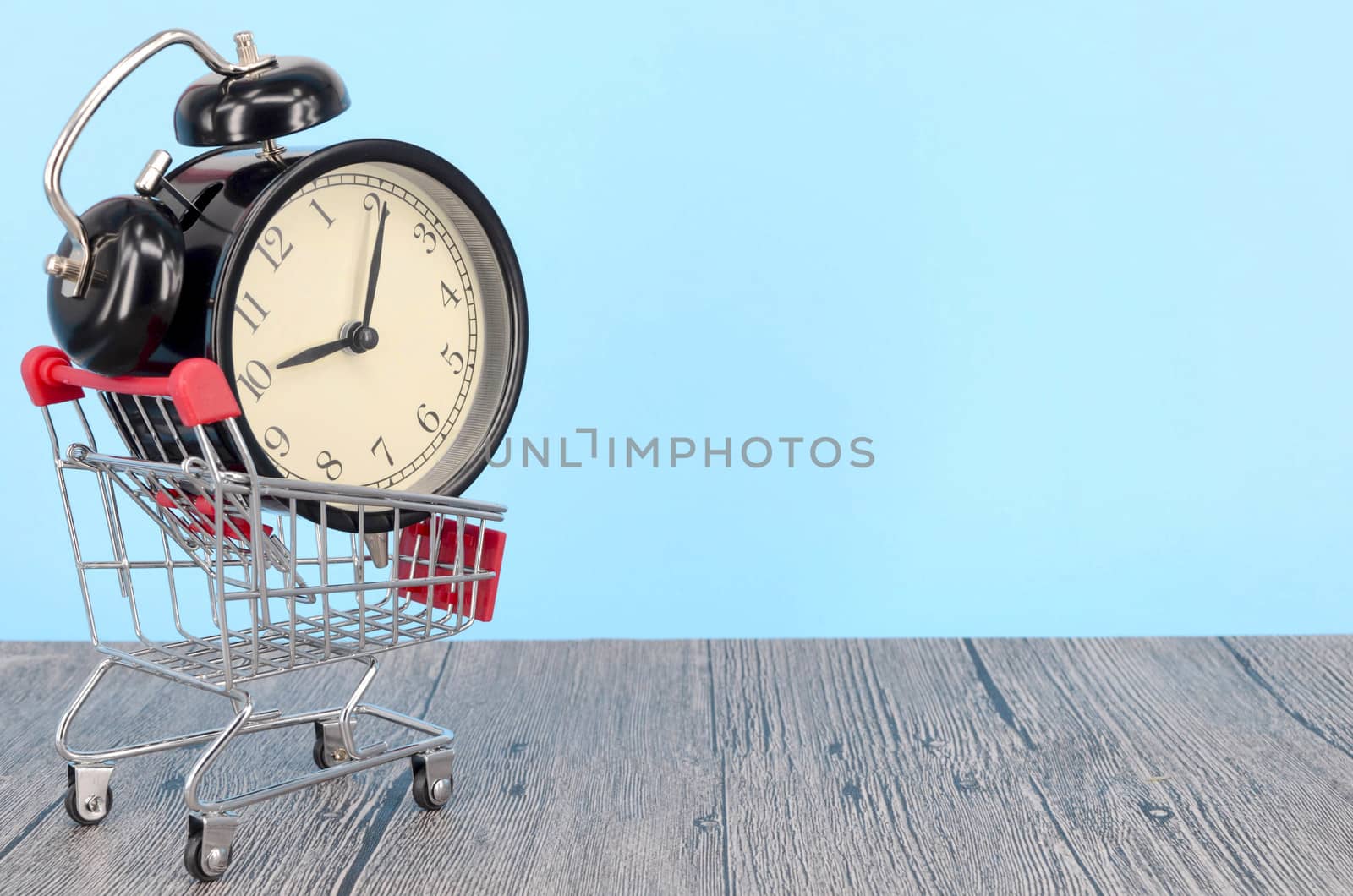 Shopping cart and classic alarm clock on wooden surface. Sale time buy mall market shop consumer concept. Selective focus.