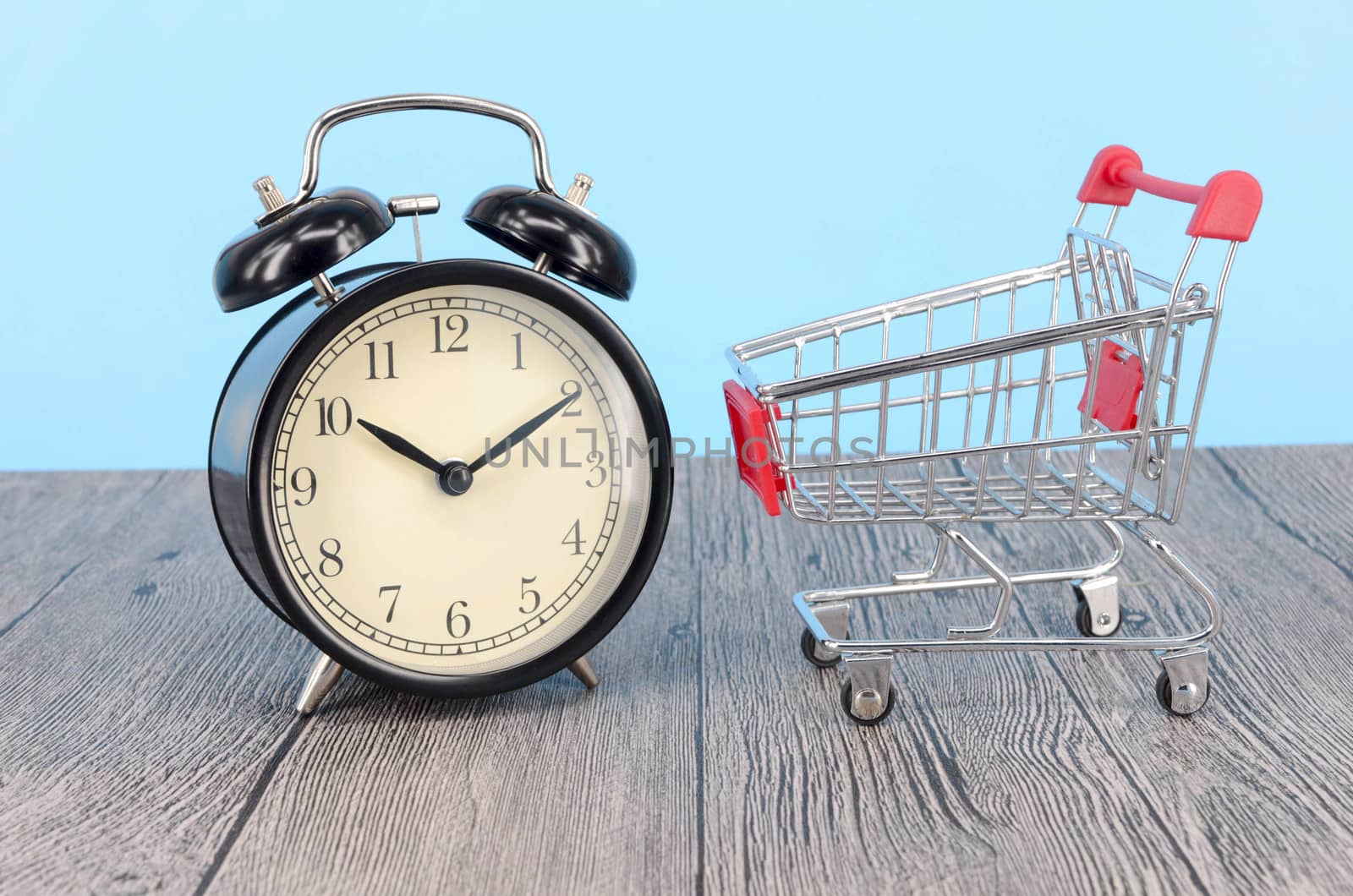 Shopping cart and classic alarm clock on wooden surface. Sale time buy mall market shop consumer concept. Selective focus.