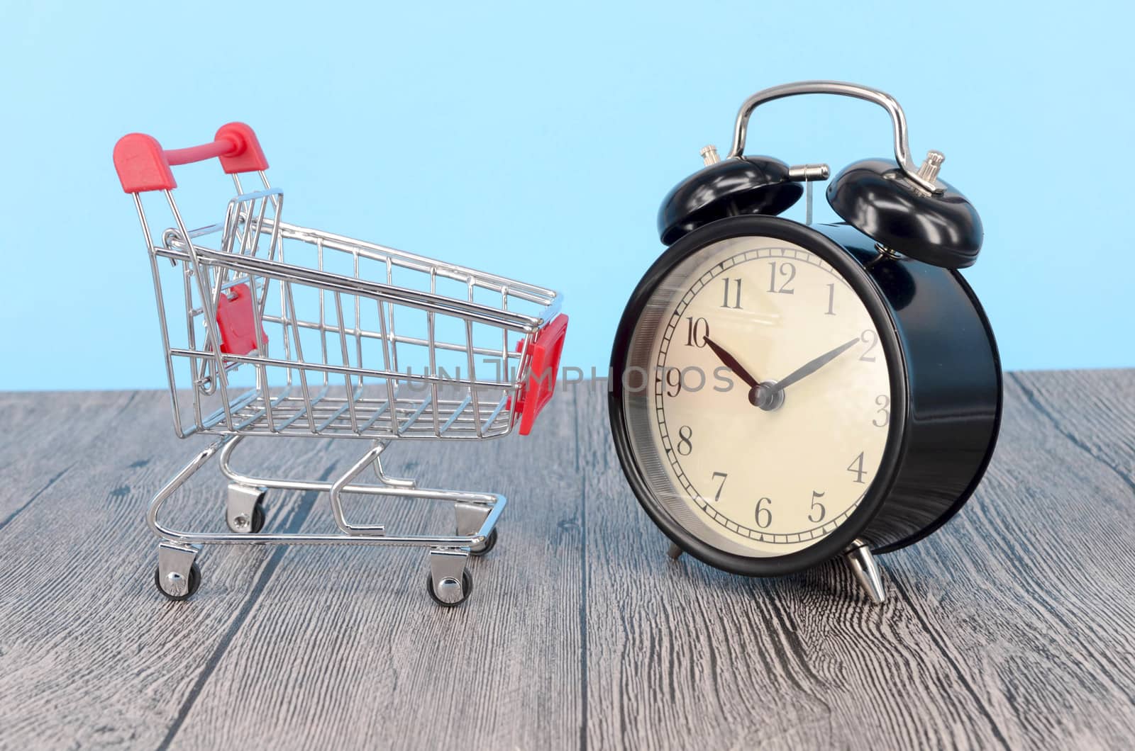 Shopping cart and classic alarm clock on wooden surface. Sale time buy mall market shop consumer concept. Selective focus.