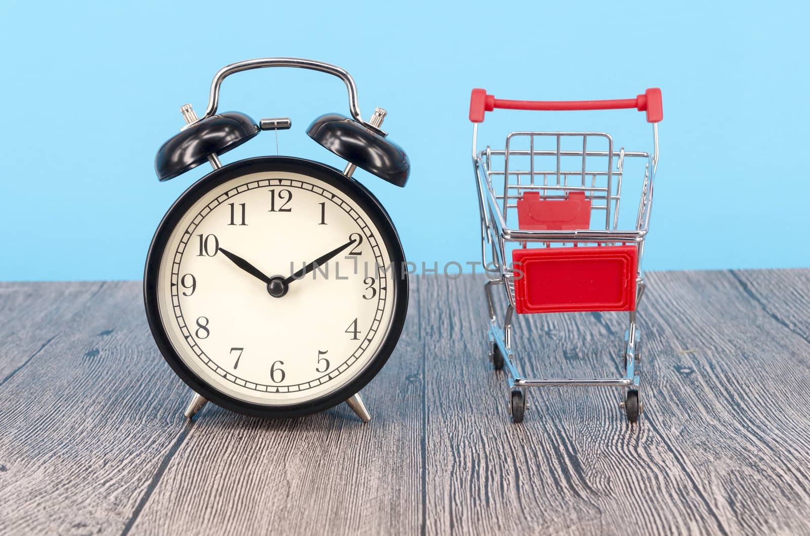 Shopping cart and classic alarm clock on wooden surface. Sale time buy mall market shop consumer concept. Selective focus.