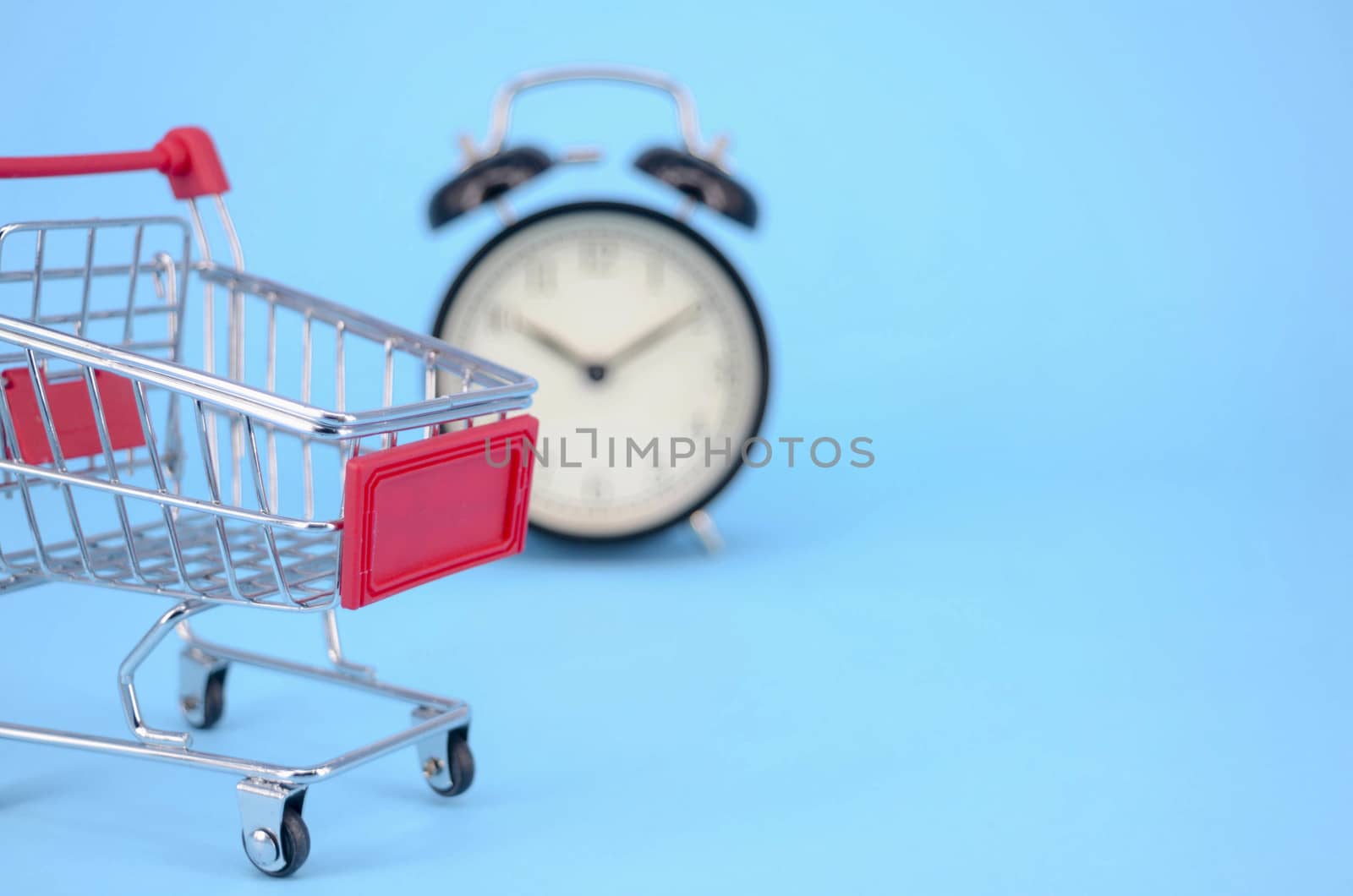 Shopping cart and classic alarm clock on blue background. Sale time market shop consumer concept. Selective focus.