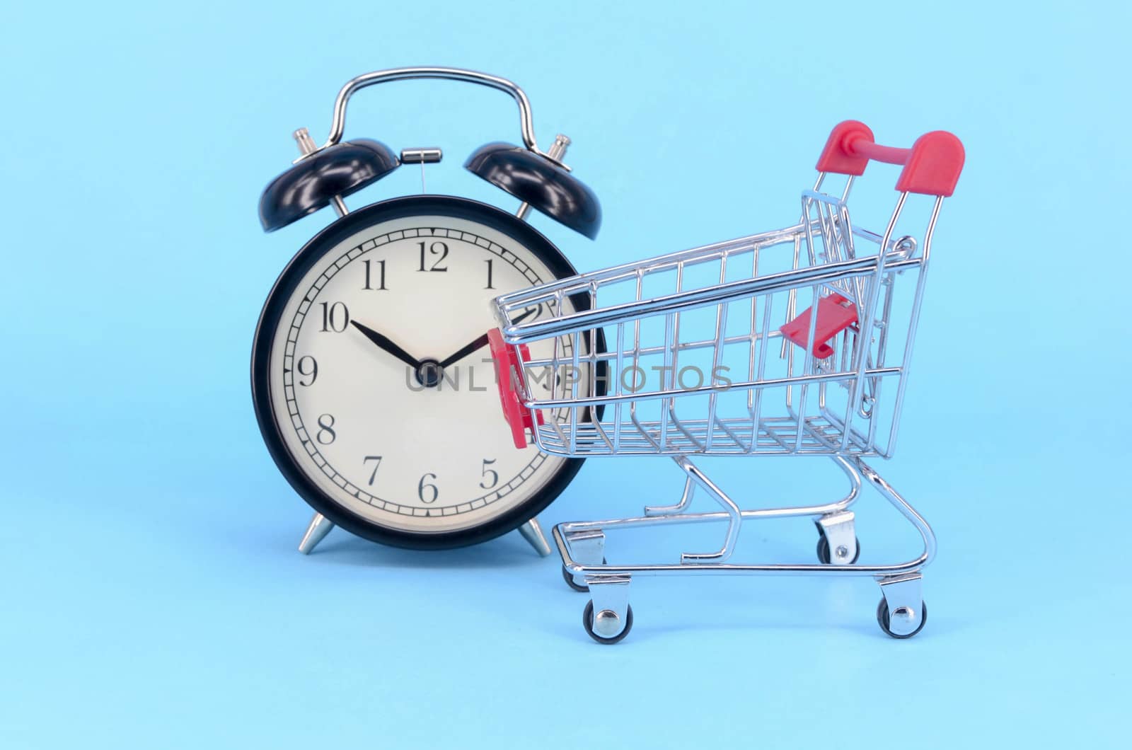 Shopping cart and classic alarm clock on blue background. Sale time market shop consumer concept. Selective focus.