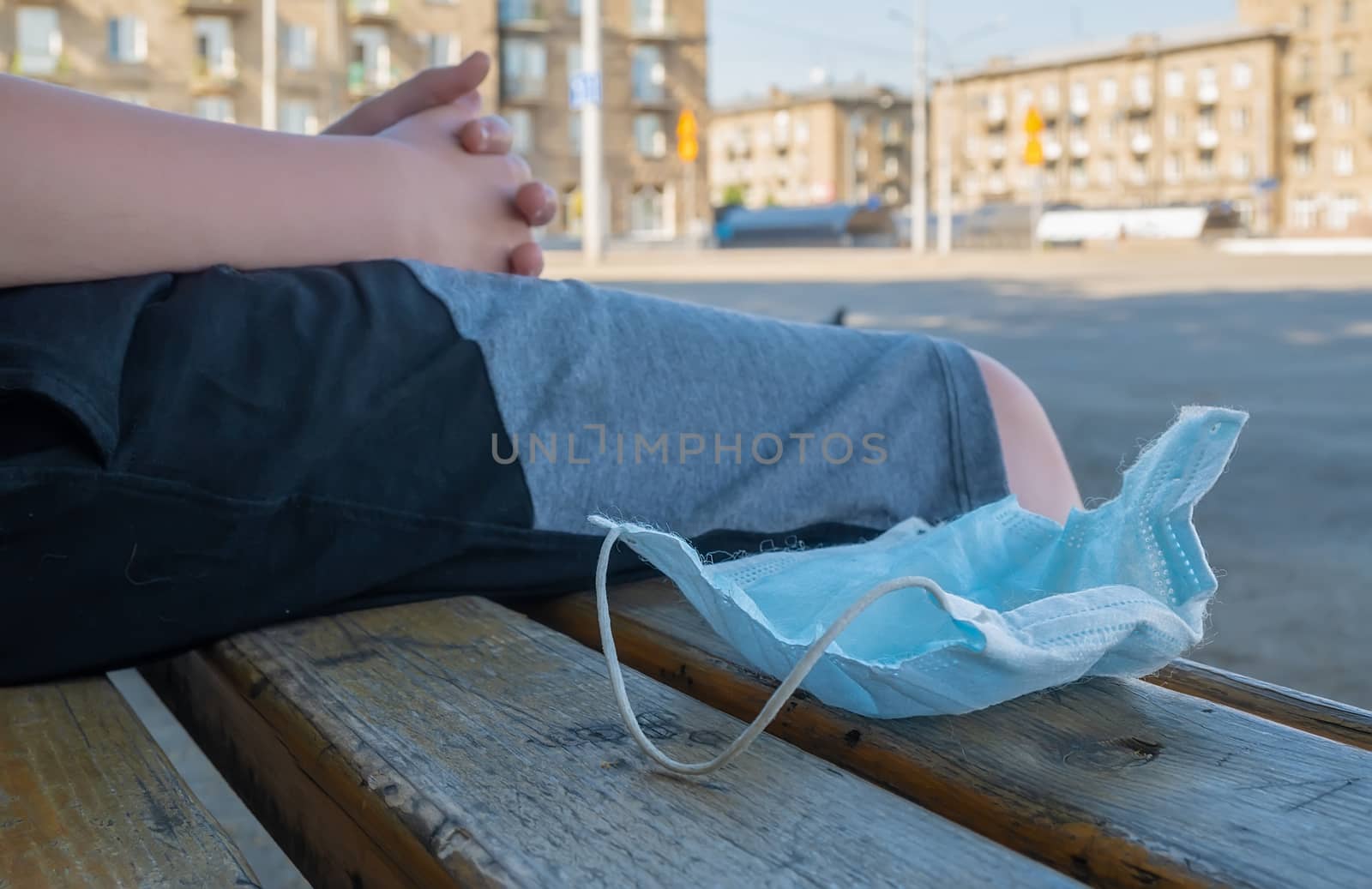 abandoned and used antibacterial medical mask lies on the asphalt at the feet of a man who is sitting on a bench in a city Park