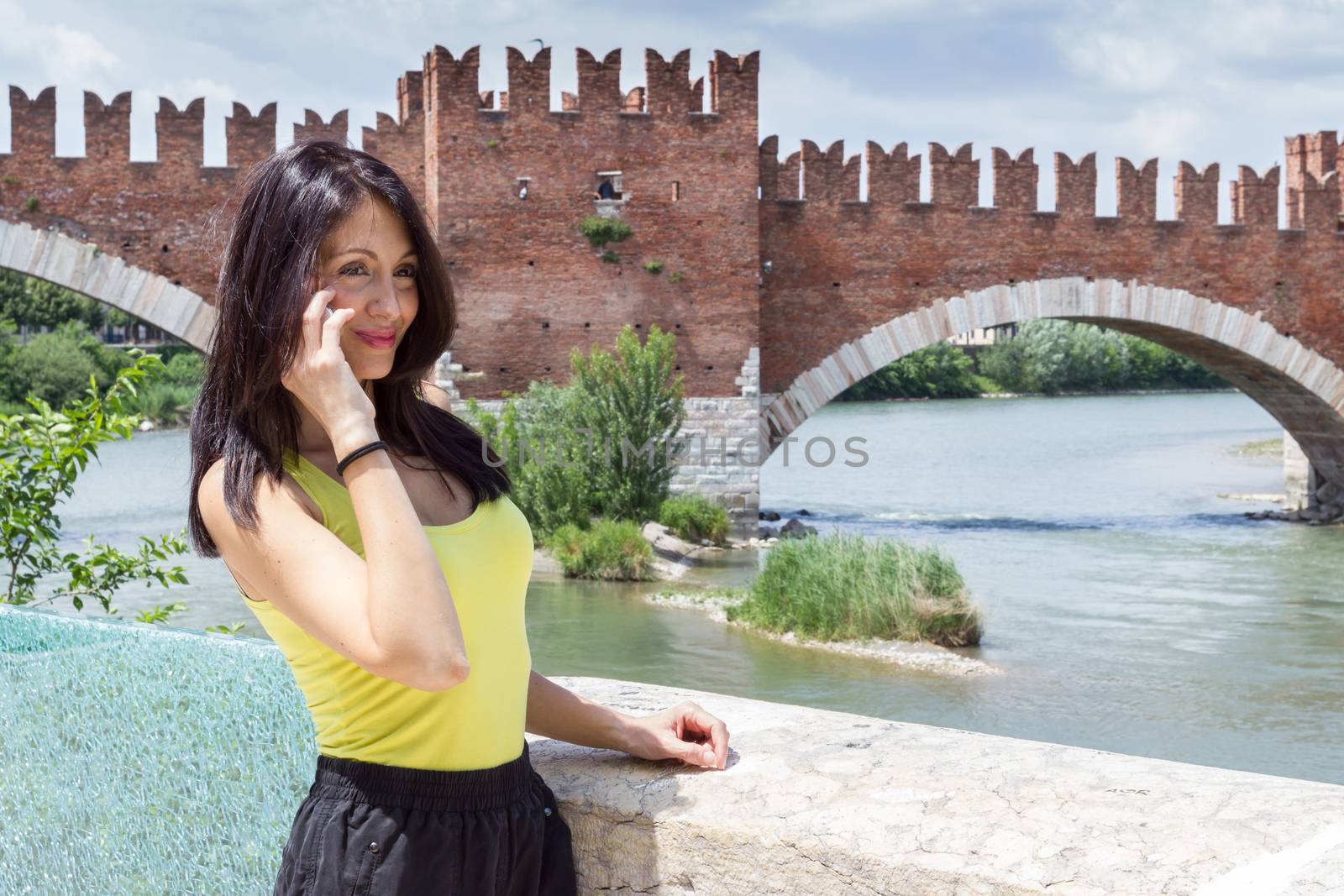 Portrait of a young woman talking on phone. In the background the medieval stone bridge Scaligero built in 14th century near Castelvecchio, Verona (Italy). Shallow DOF.