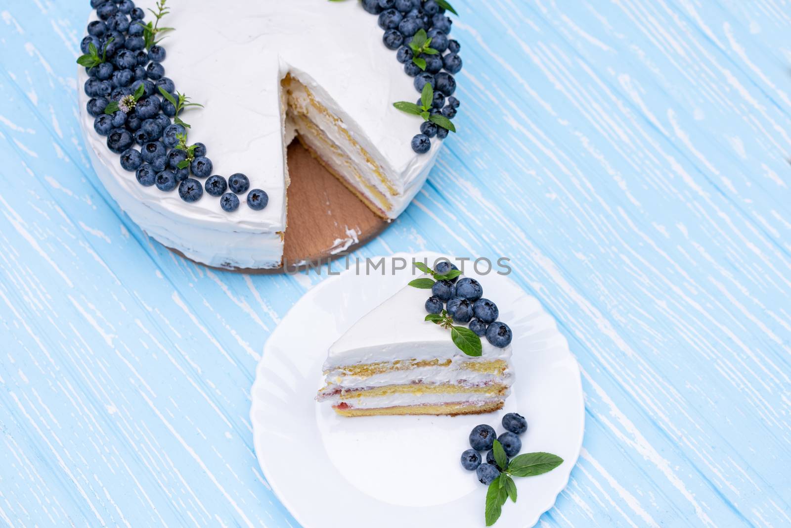 Appetizing cheesecake biscuit pillow decorated white cream blueberries and mint stands on wooden blue rustic table. Sweet cake with piece on plate