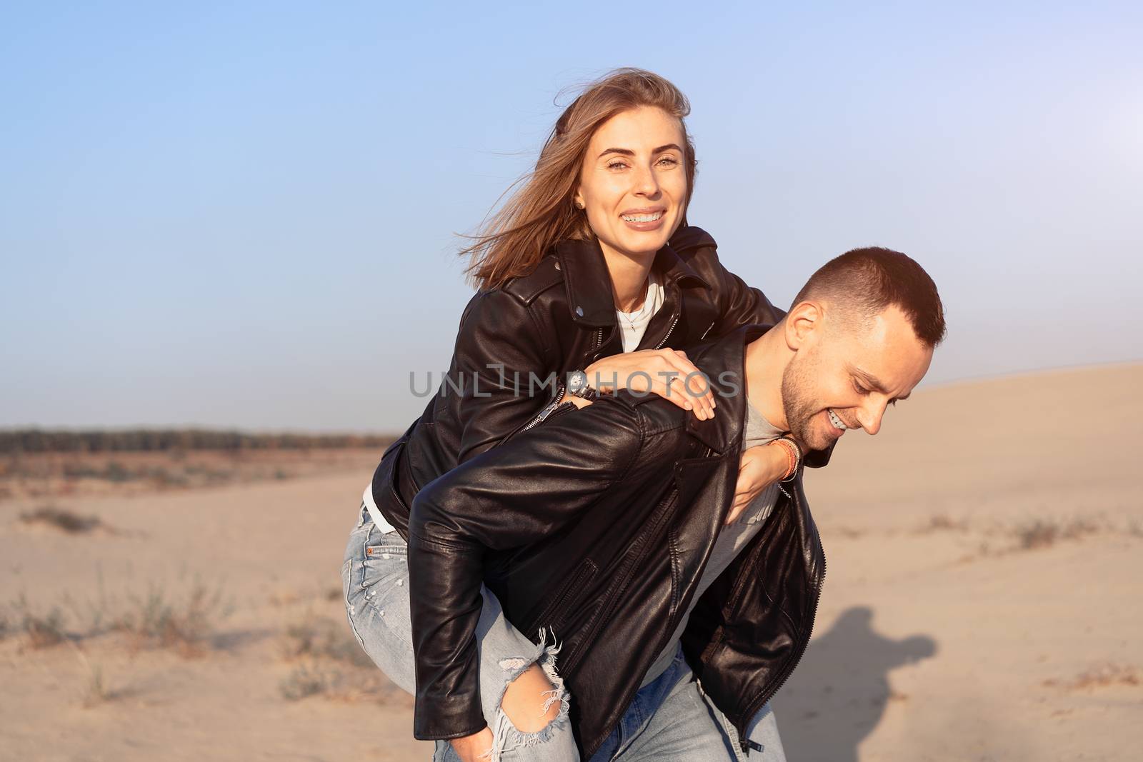 Happy and cute adorable adult couple leather jacket and jeans man with woman girlfriend on piggy back, have fun play, laugh,smile and jump on sunset at desert crazy in love, emotions and relationship
