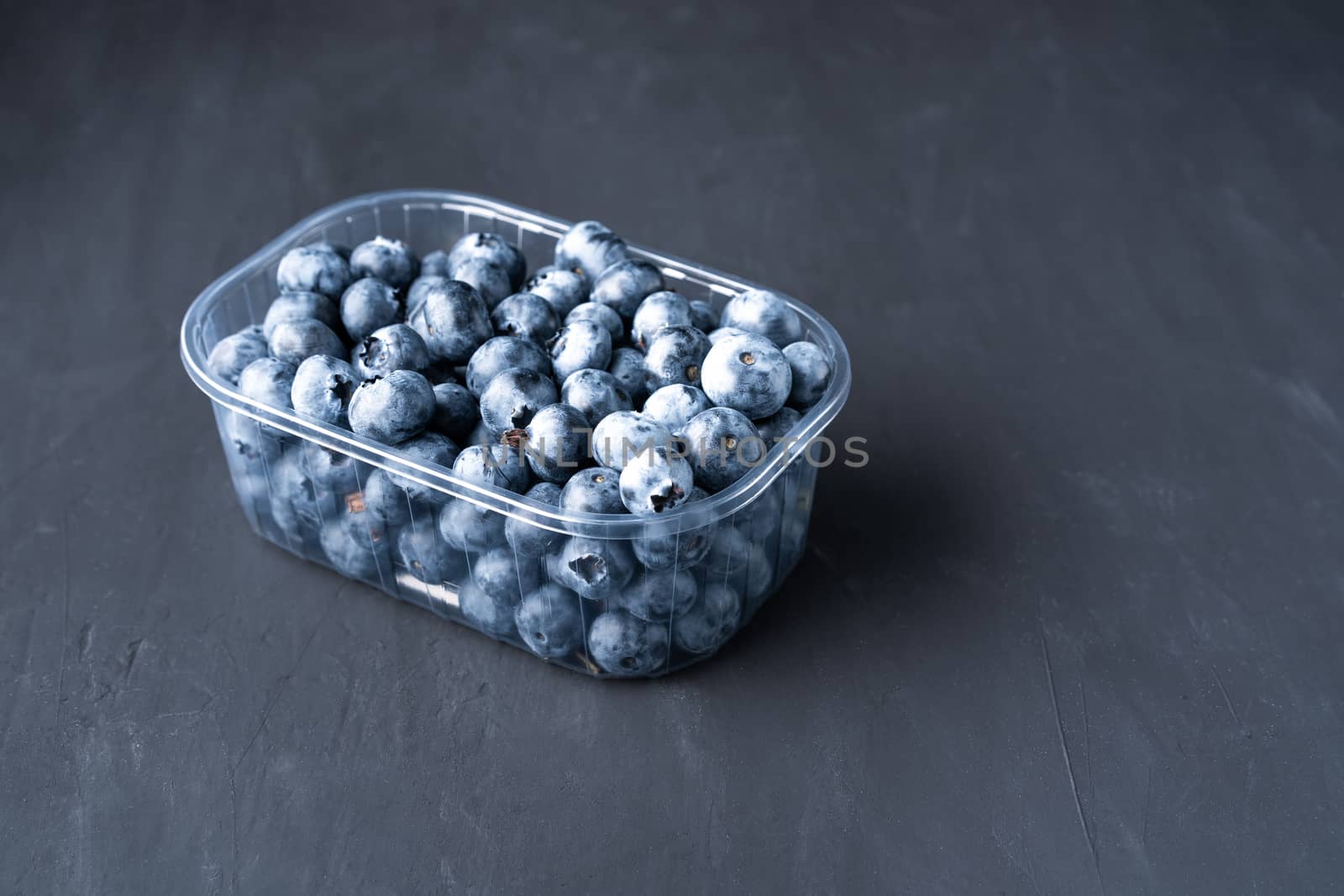 Tasty juicy raw blueberries in a plastic container on a black dark background. Packaging for berries in a supermarket on store shelves.