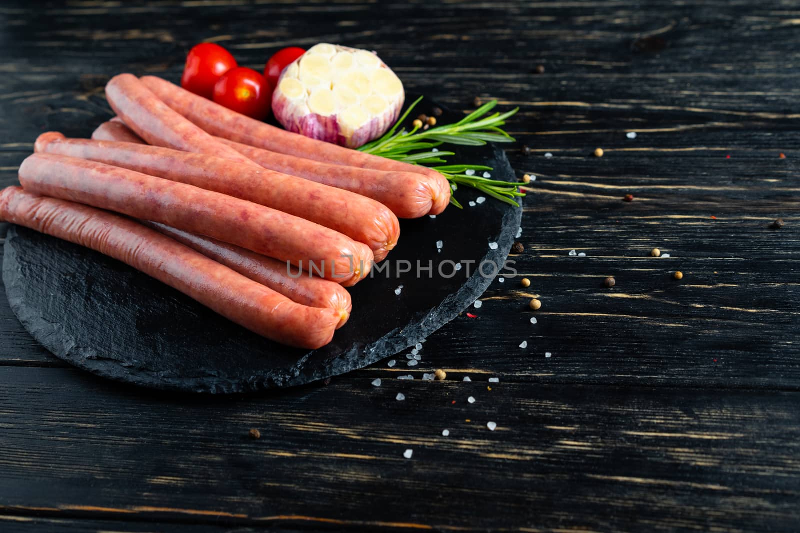 Juicy sausages for barbecue lie on a stone chopping board with rosemary garlic cherry tomatoes spices, pepper and coarse salt on the black wooden table background