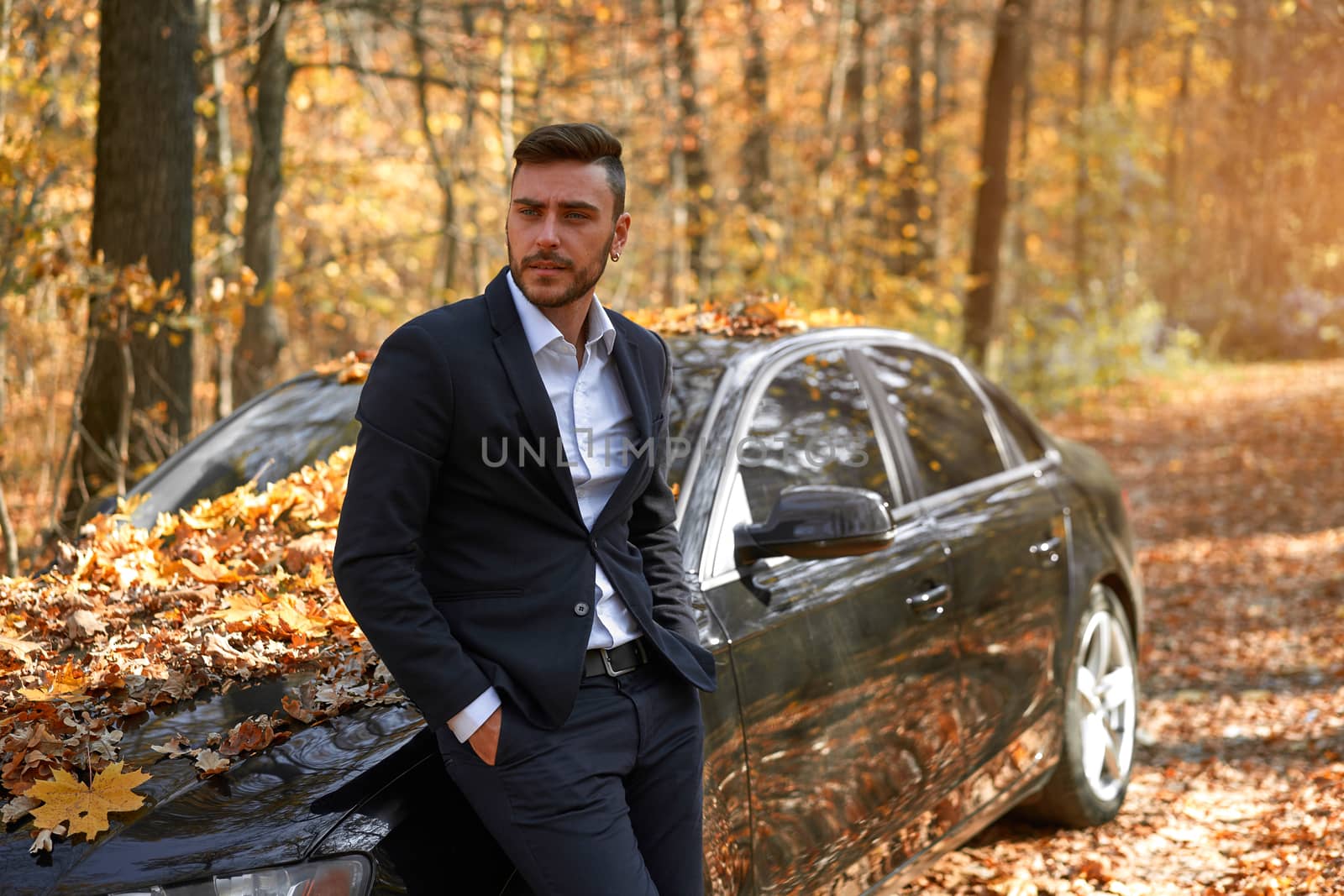 A handsome young Caucasian man wearing business suit standing near black car on nature. by andreonegin