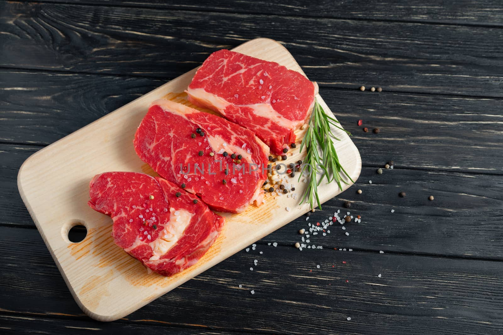 Three pieces of juicy raw beef with rosemary on a cutting board on a black wooden table background. Meat for barbecue or grill sprinkled with pepper and salt seasoning