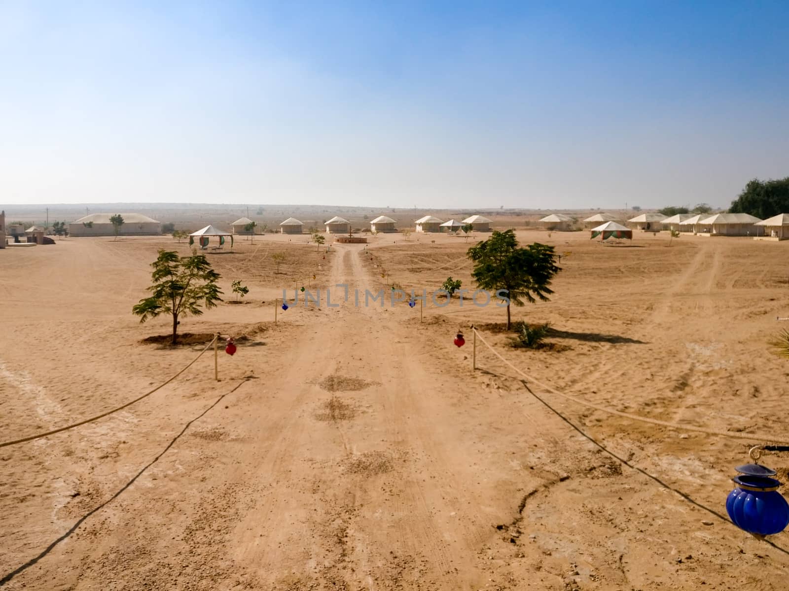 Empty dirt road with barren desert on both sides and blue sky above showing thar desert of rajasthan india. Shows the tents of a desert camp in the distance with lanterns hanging on the side