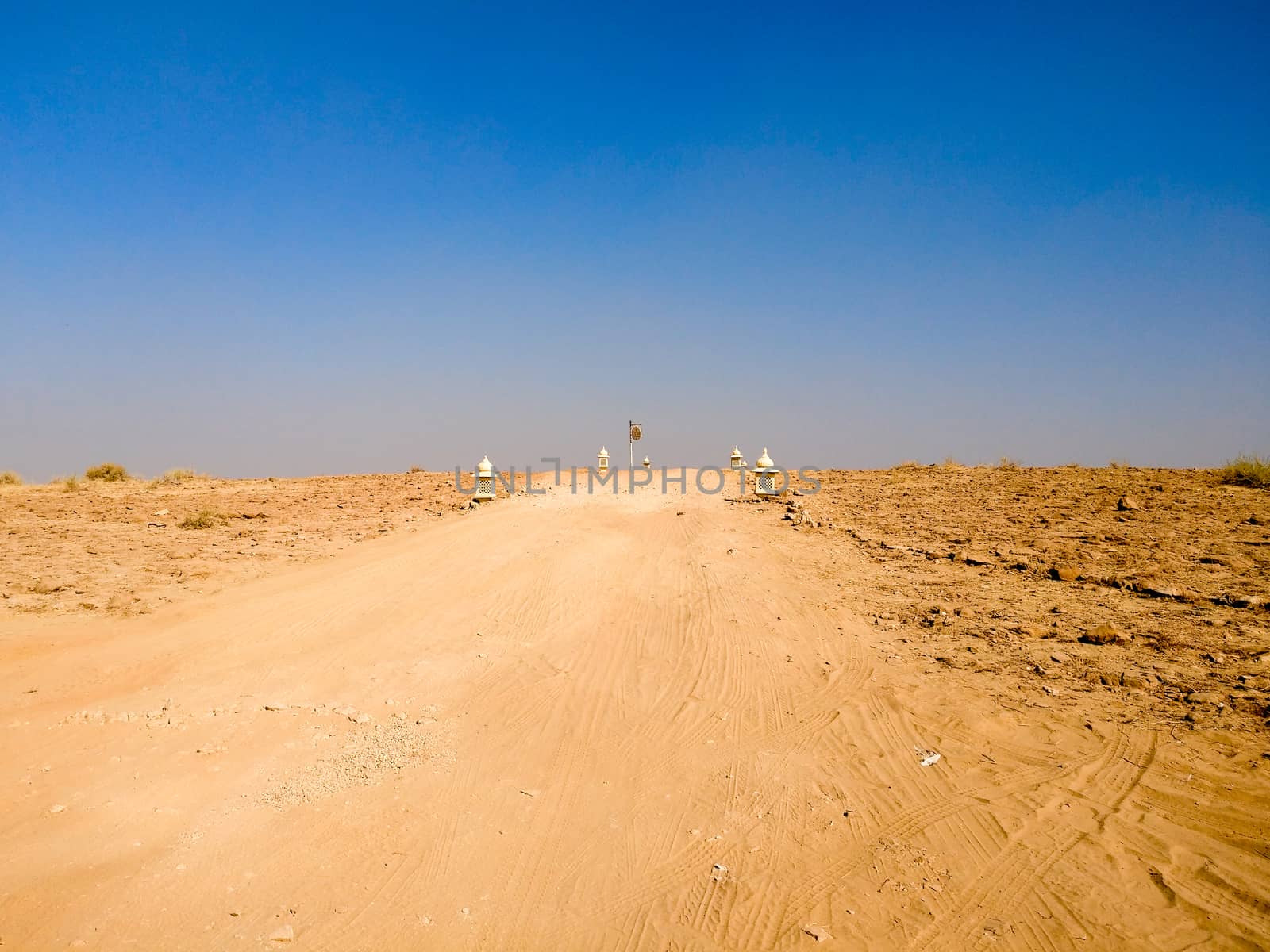 Empty dirt road with barren desert on both sides and blue sky above showing thar desert of rajasthan india by Shalinimathur