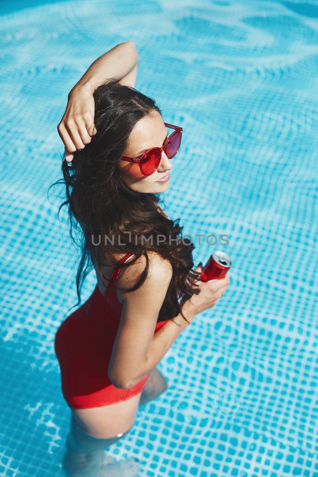 Fashionable and happy brunette model woman with a perfect sexy figure in stylish red swimsuit and trendy red sunglasses in the swimming pool holding a cold drink in her hand.