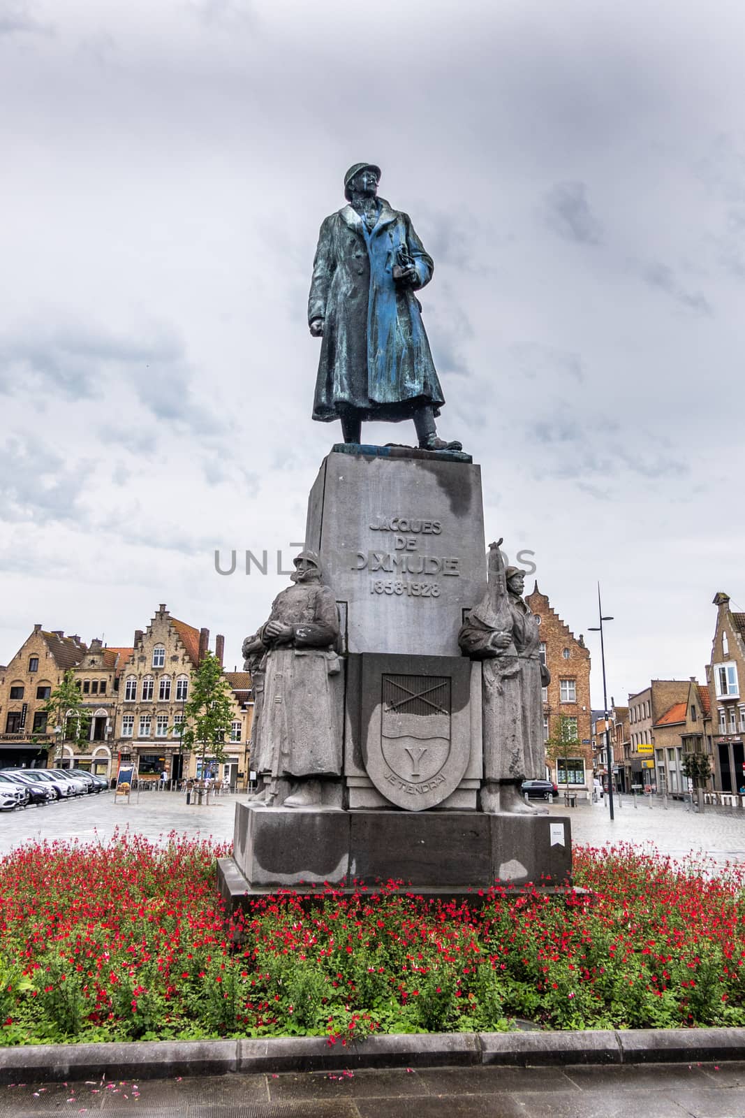 Diksmuide, Flanders, Belgium -  June 19, 2019: Grote Markt. Bronze statue of General Jacques de Dixmude on gray stone pedestal in red flowers against silver sky. Some Yellowish brown house facades.