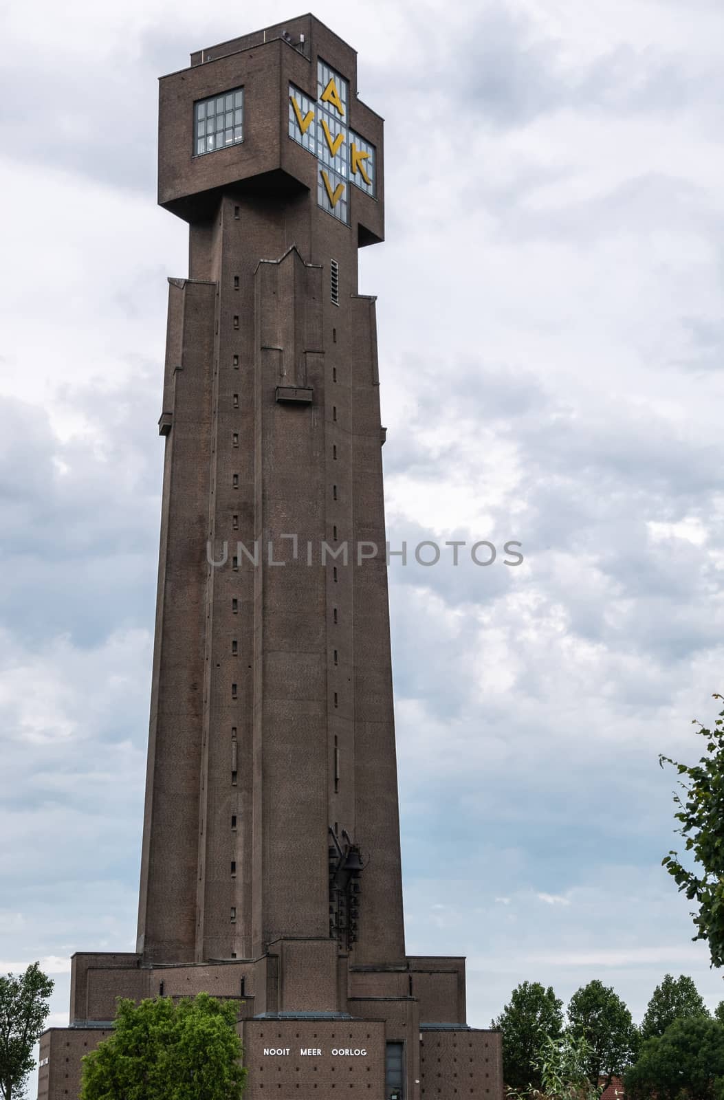 Diksmuide, Flanders, Belgium -  June 19, 2019: Closeup of IJzertoren, tallest peace monument of WW 1 against gray blue cloudscape. Dutch words saying No More War. Some green foliage.