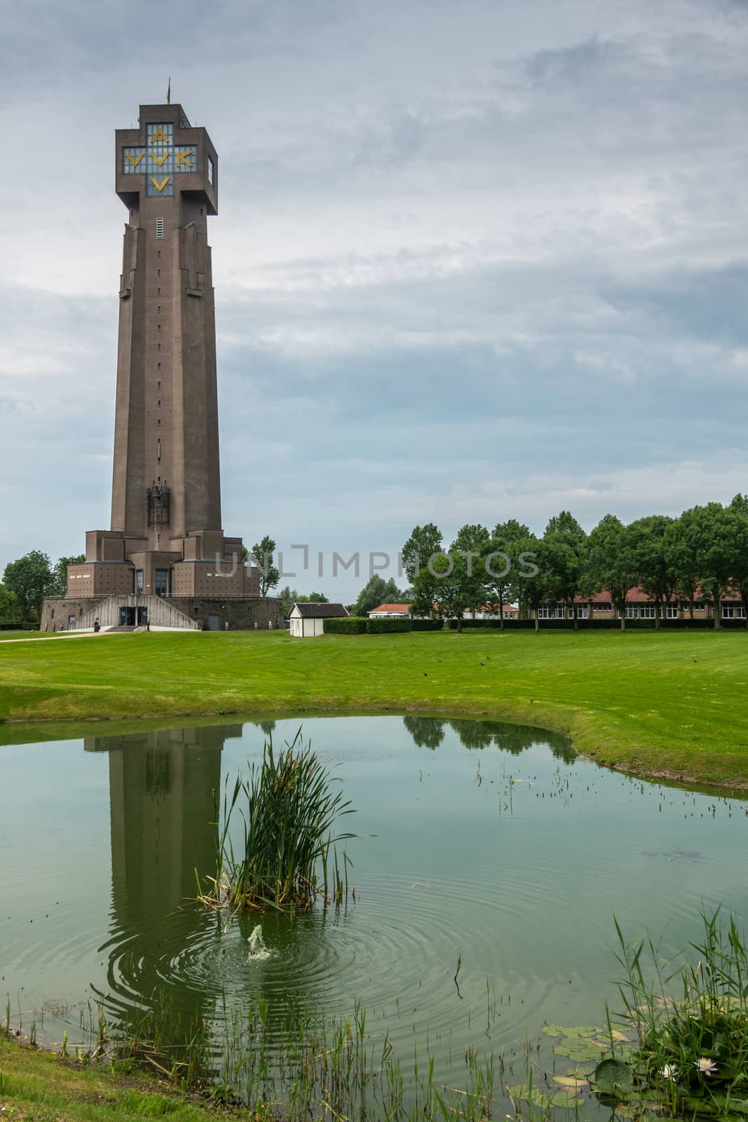 Diksmuide, Flanders, Belgium -  June 19, 2019: IJzertoren, tallest peace monument of WW 1 against gray blue cloudscape. On wall saying No More War. Reflection in pond. Some green foliage and lawn.