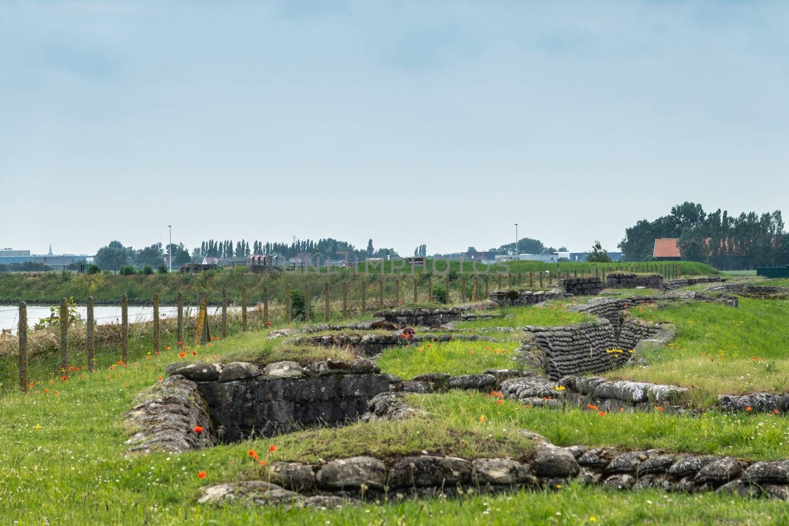 Diksmuide, Flanders, Belgium -  June 19, 2019: Overview of Historic WW1 Trenches, called Dodengang along IJzer River, shows gray-brown stone-hard sandbags, green grass, red poppies. Barbed wire fence and river under blue sky.