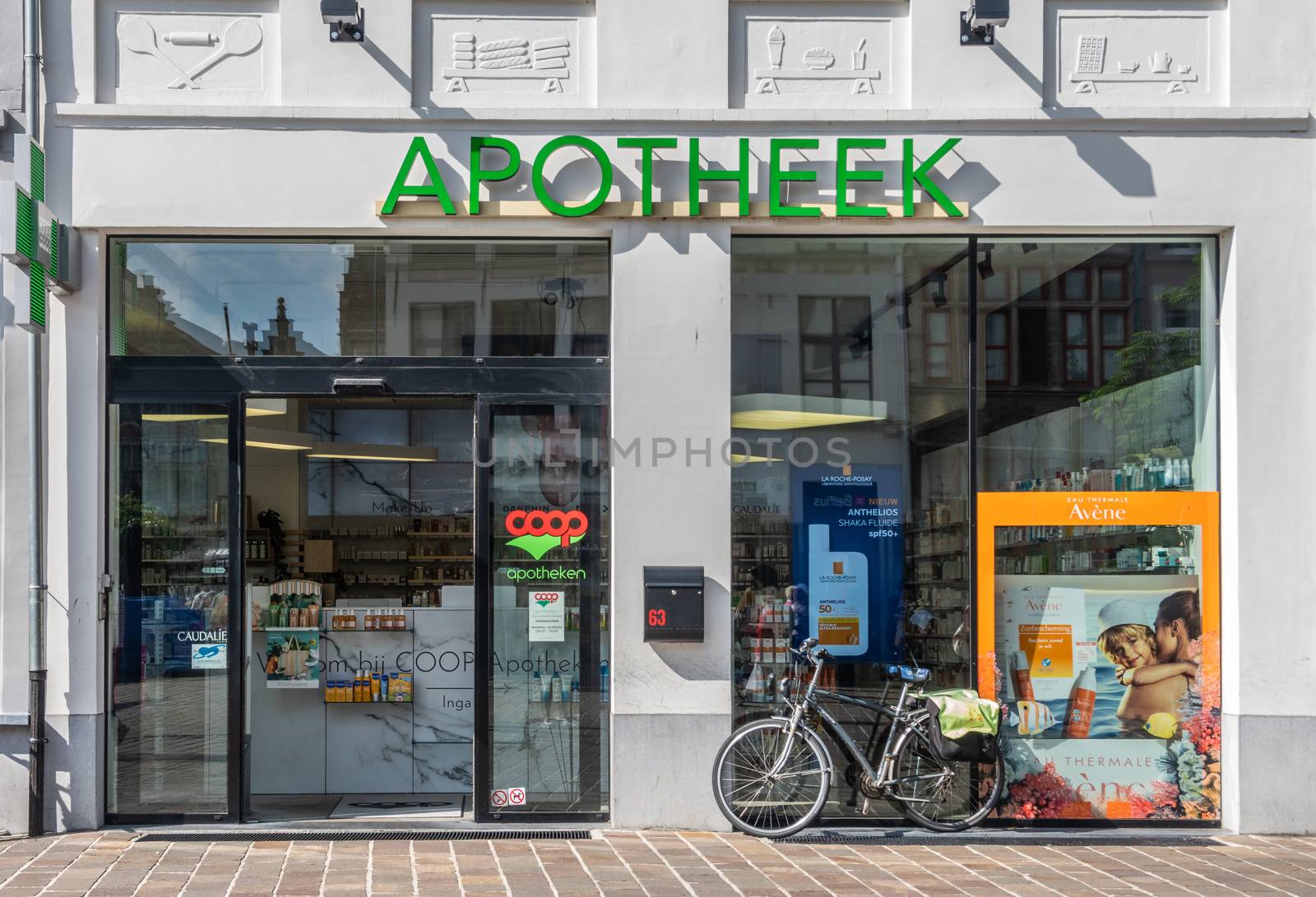 Gent, Flanders, Belgium -  June 21, 2019: Front large windows of COOP Socialist Pharmacy in Langemunt street, set in old bakery building as gray wall frescoes show. Drug advertisements and bike.