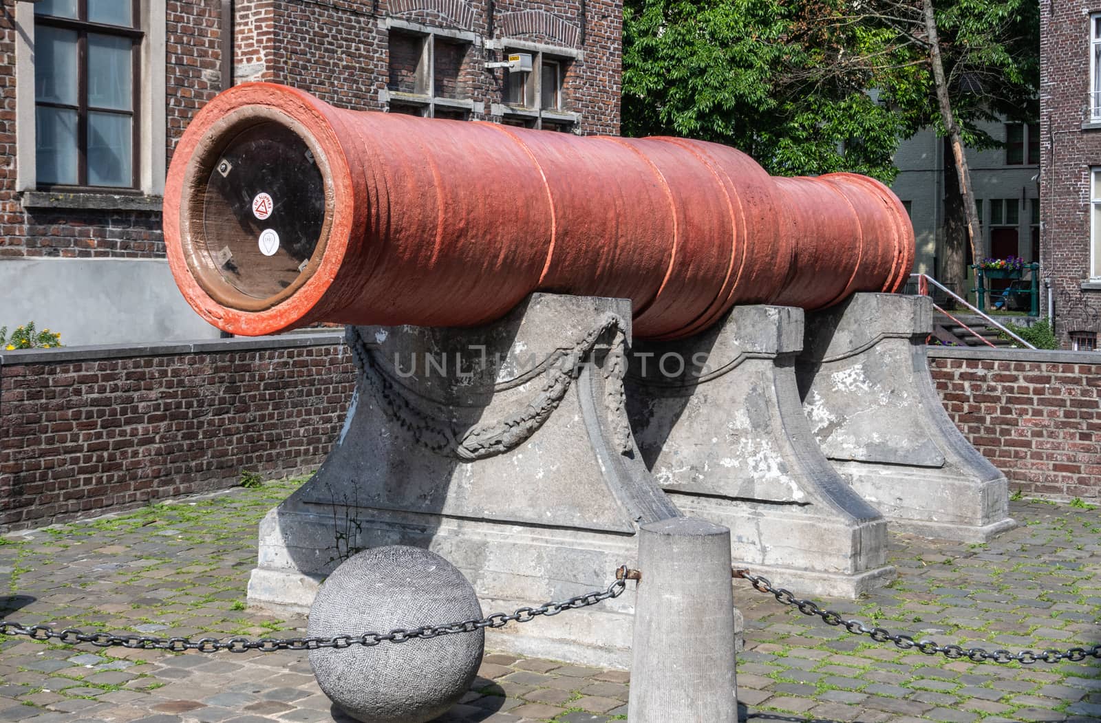 Gent, Flanders, Belgium -  June 21, 2019: Historic large, red, Dulle Griet Cannon with stone cannon ball. Brick wall with window and green foliage as background.