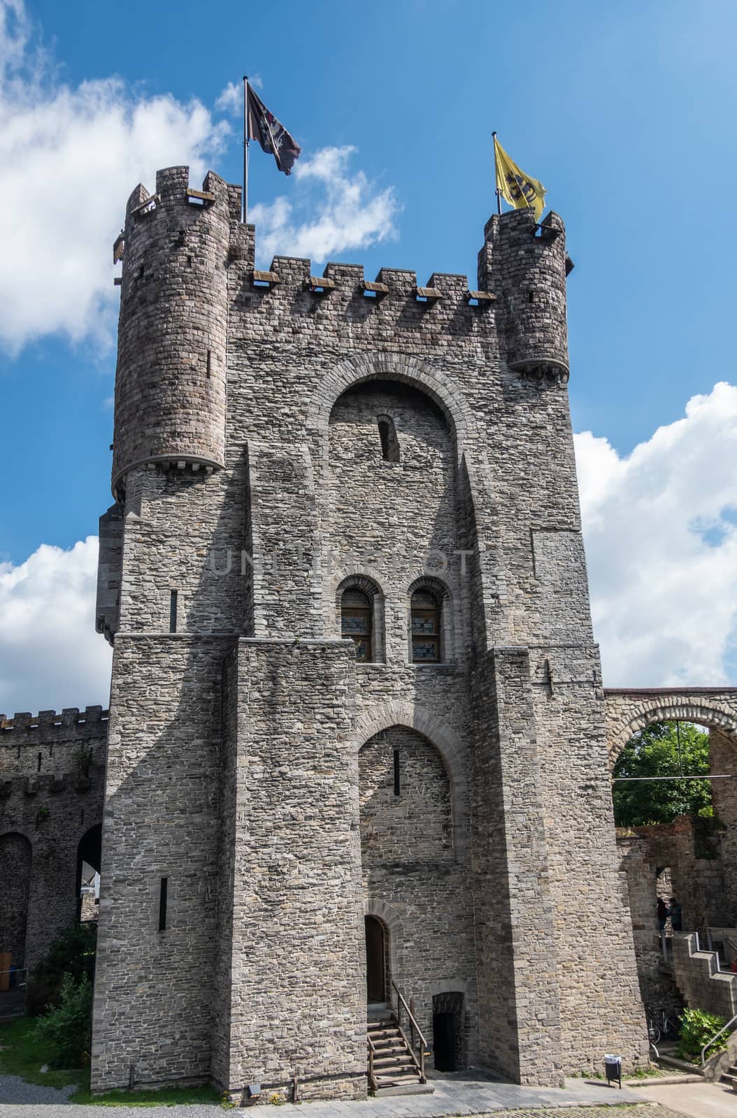 Gent, Flanders, Belgium -  June 21, 2019: Gray stone tower of Gravensteen, historic medieval castle of city against blue sky with white clouds. Flags on top, Green foliage.