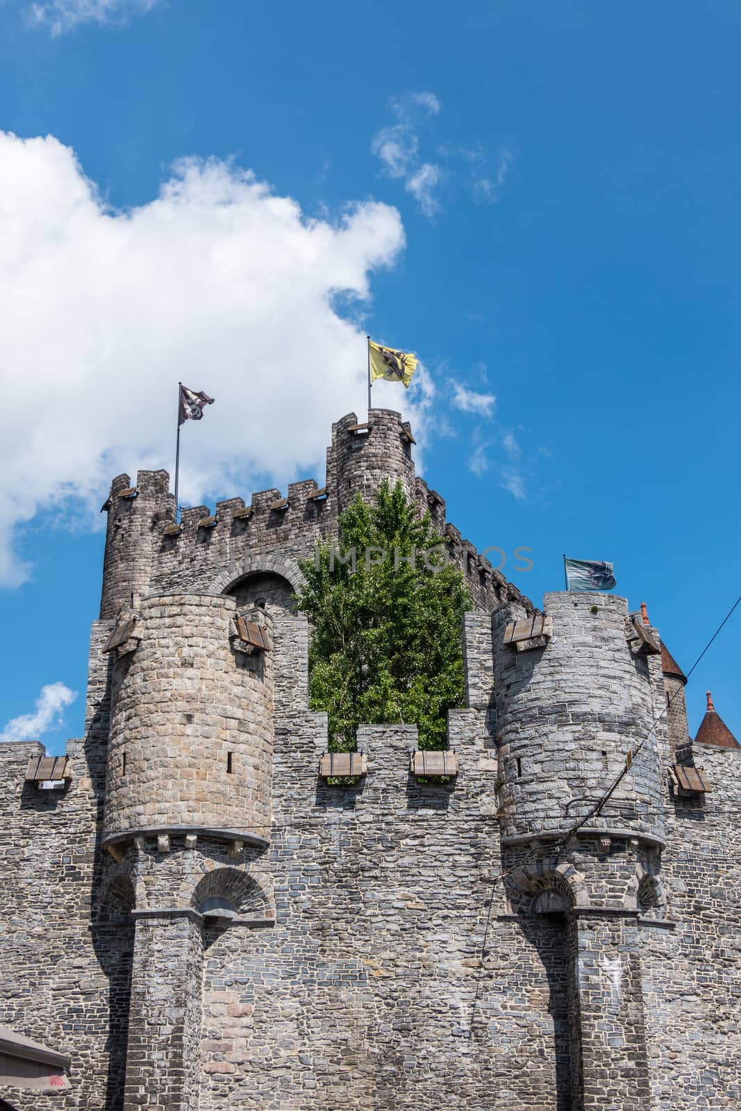 Gent, Flanders, Belgium -  June 21, 2019: Gray stone tower and ramparts of Gravensteen, historic medieval castle of city against blue sky with white clouds. Flags on top, green foliage.