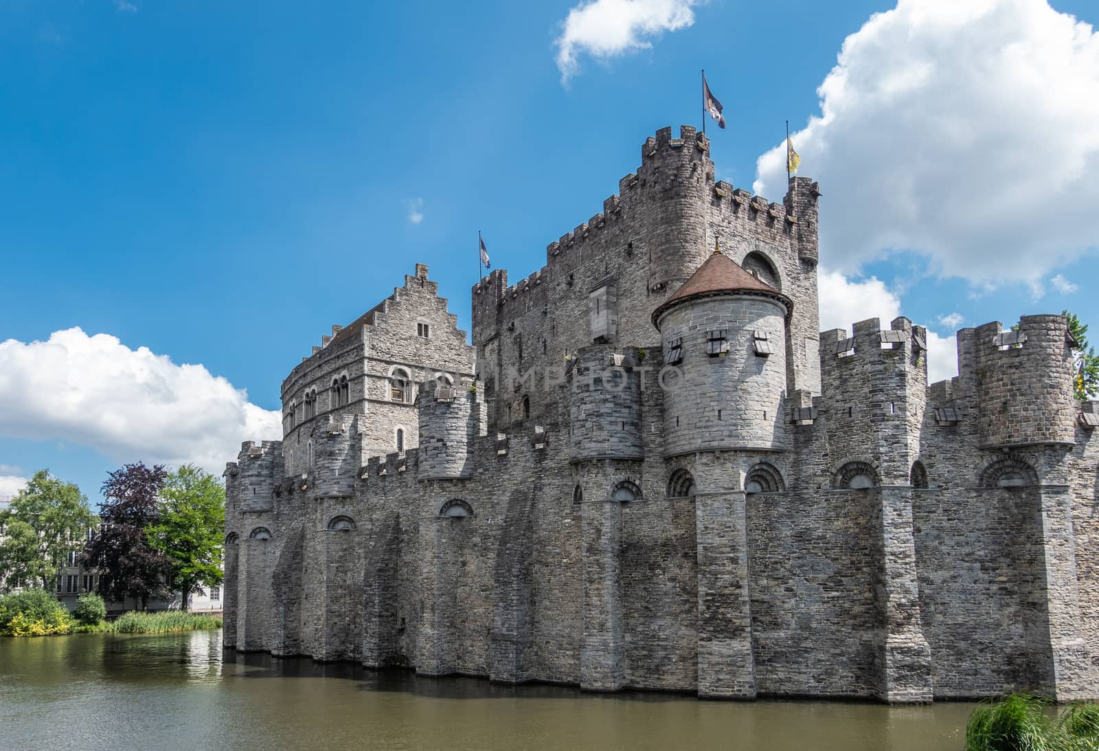 Gent, Flanders, Belgium -  June 21, 2019: Gray stone cast;e and ramparts of Gravensteen, historic medieval castle of city, behind its moad against blue sky with white clouds. Flags on top, green foliage.