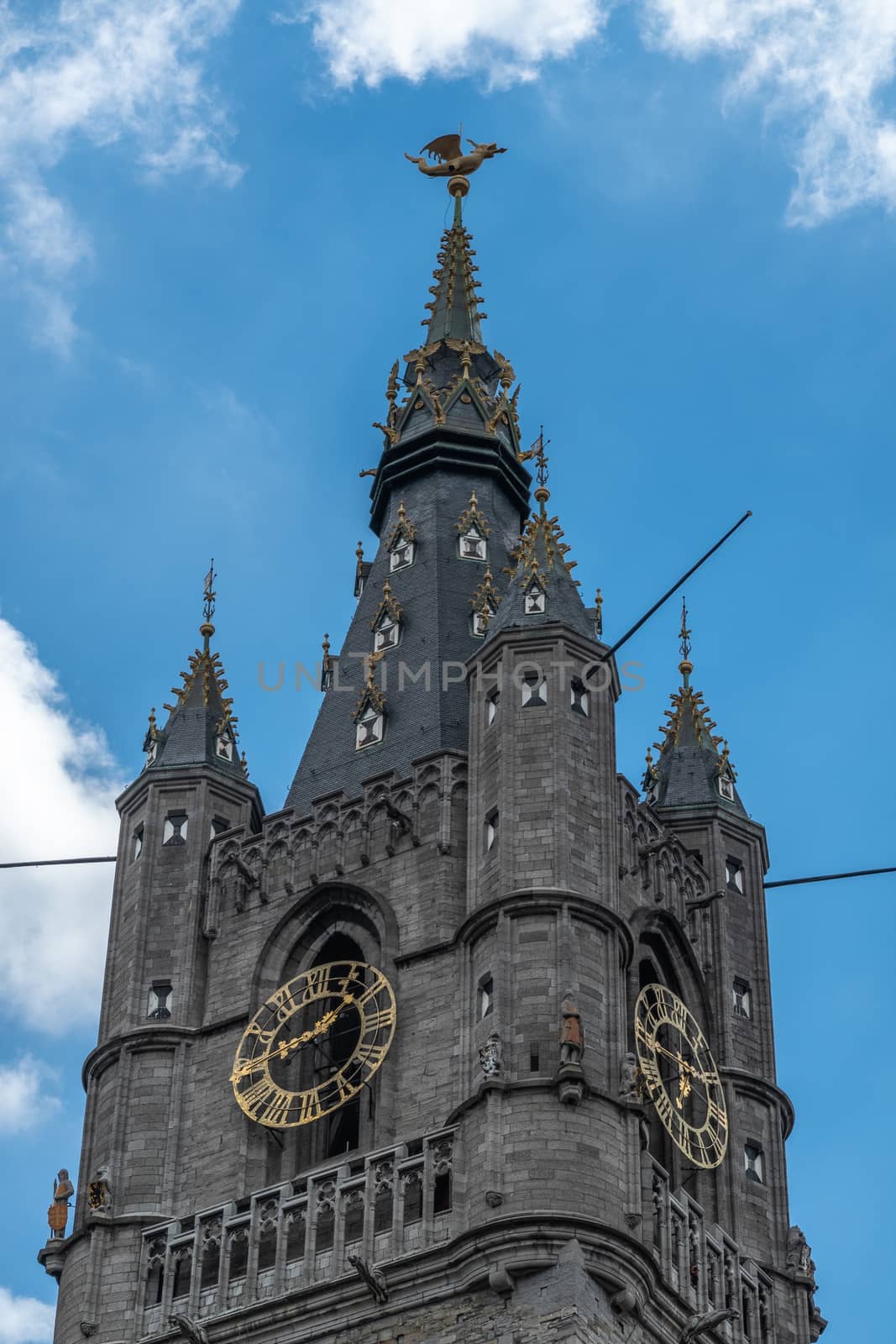 Gent, Flanders, Belgium -  June 21, 2019: Closeup of top of dark stone Belfry clock tower against blue sky with white patches. Gulden Draak, dragon on pinacle.