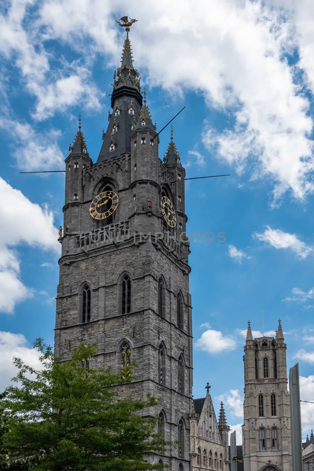 Gent, Flanders, Belgium -  June 21, 2019: Belfry clock tower with Gulden Draak dragon statue and Cathedral tower in back, all against blue sky with white clouds. Green foliage.
