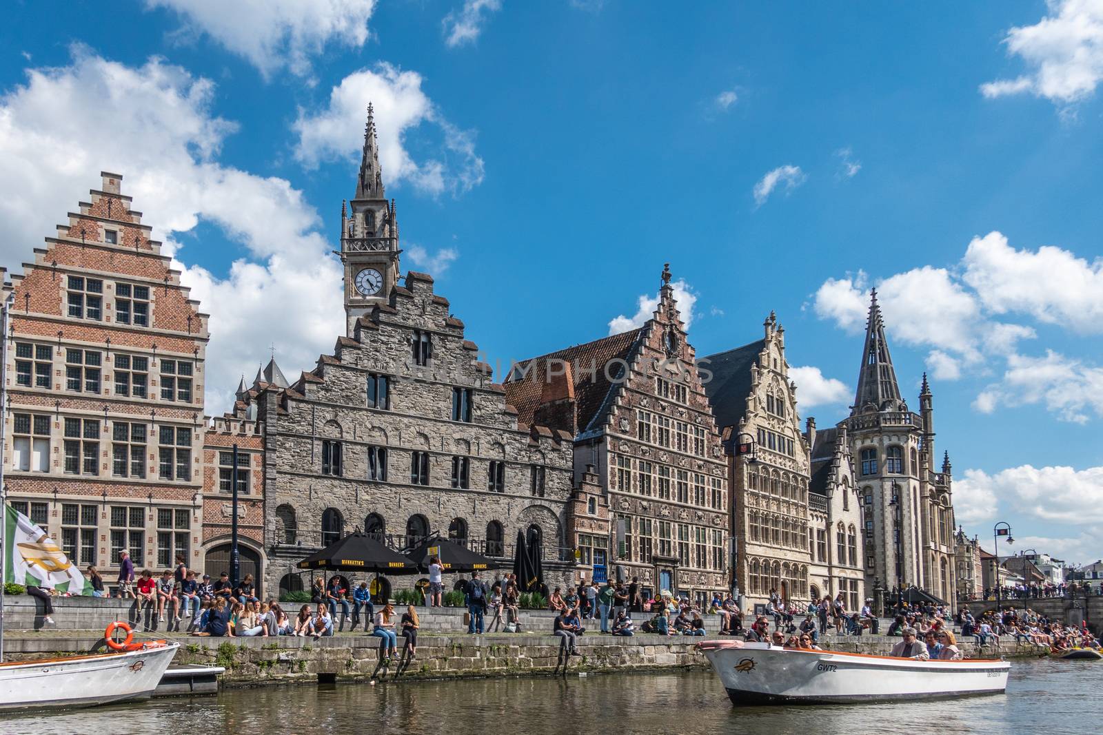 Gent, Flanders, Belgium -  June 21, 2019: School children and students enjoying sun sitting on quay of Leie River and Graslei. Row of mediavel historic buildings. Boats, blue sky and clouds.