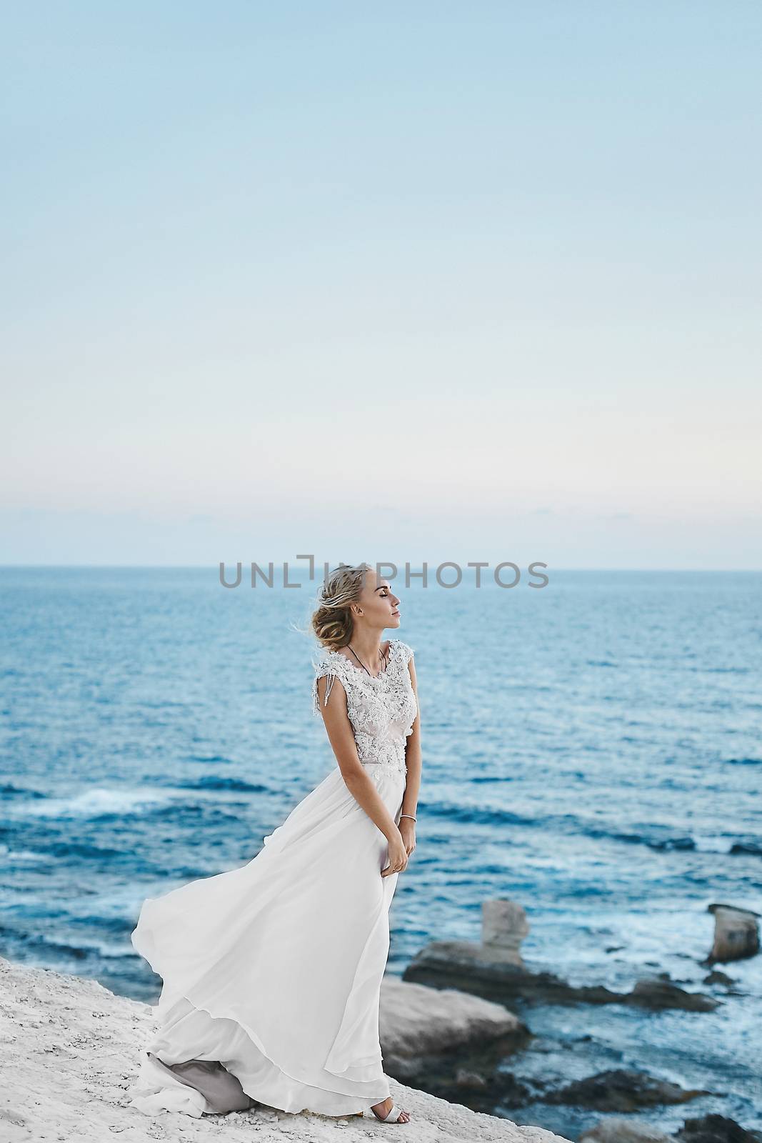 Sensual and fashionable blonde model girl with perfect body in stylish white lace dress with naked back, stands on the white rock at the sea coast.