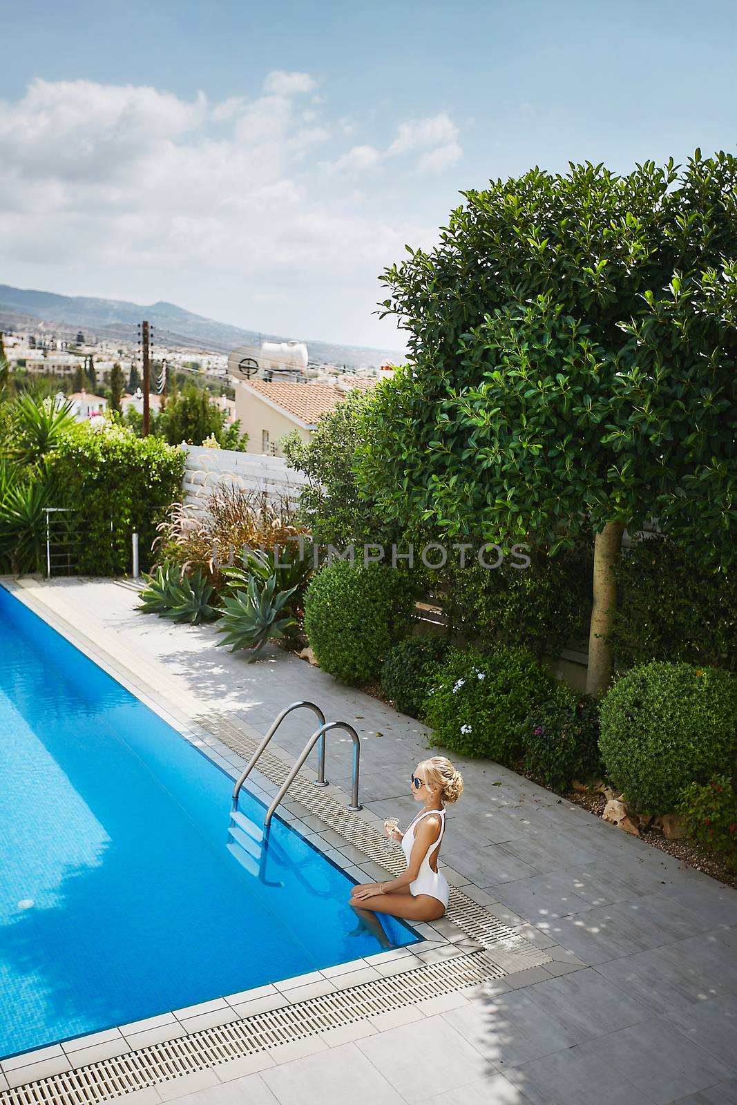Young and sexy blonde model woman with perfect body in stylish white swimsuit and in sunglasses holding a glass of champagne and sits in the swimming pool on the open air in Cyprus.