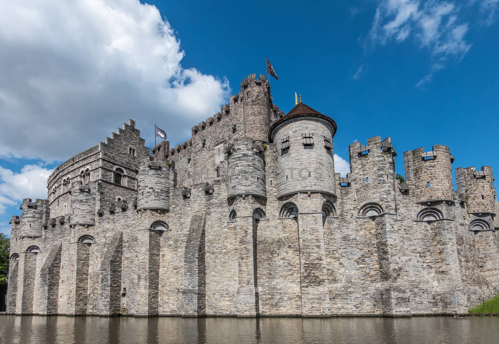Gent, Flanders, Belgium -  June 21, 2019: Gray stone castle and ramparts of Gravensteen, historic medieval castle of city, behind its moad against blue sky with white clouds. Flags on top.