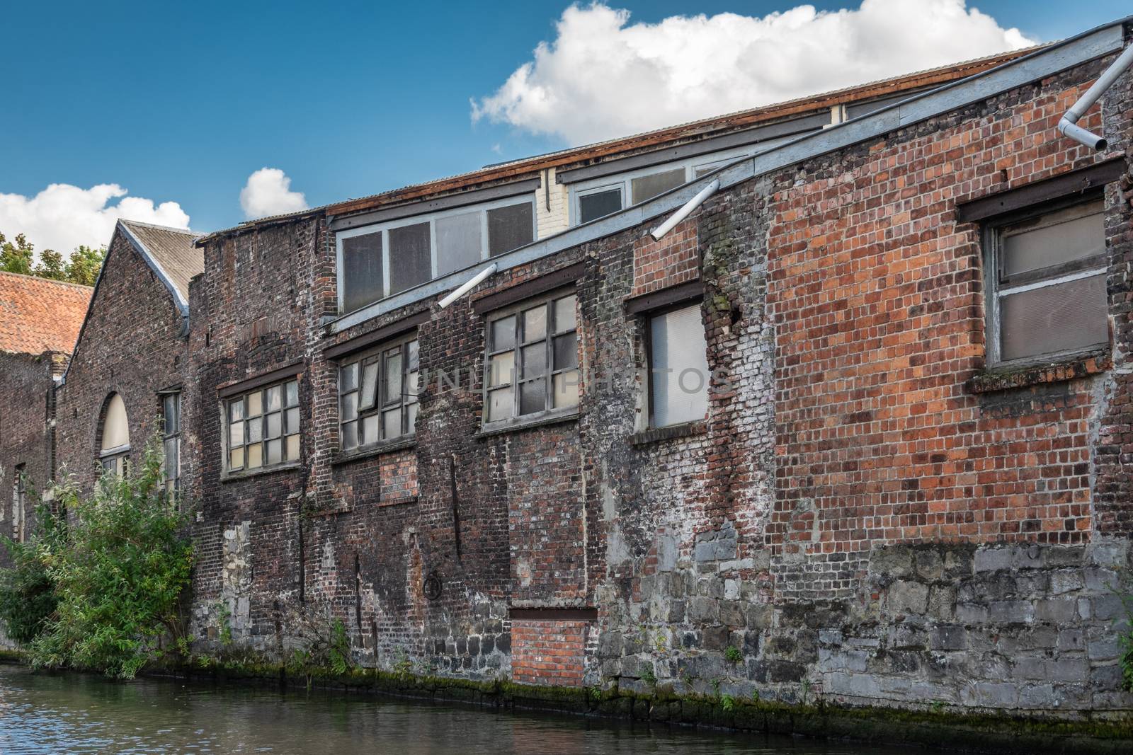 Gent, Flanders, Belgium -  June 21, 2019: Old red-brown brick industrial warehouses along the Lieve River under blue sky with white cloud patches. Greenish water.