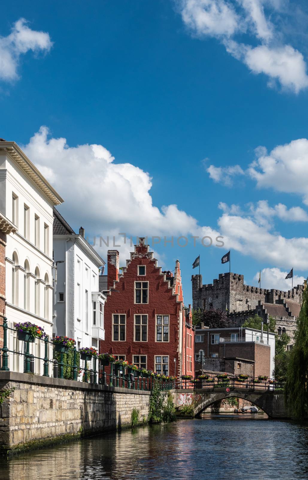 Gent, Flanders, Belgium -  June 21, 2019: Narrow Lieve River leads along old bourgeois mansions to Gravensteen Castle under blue sky with white clouds. Bluish water. Flowers on quays.