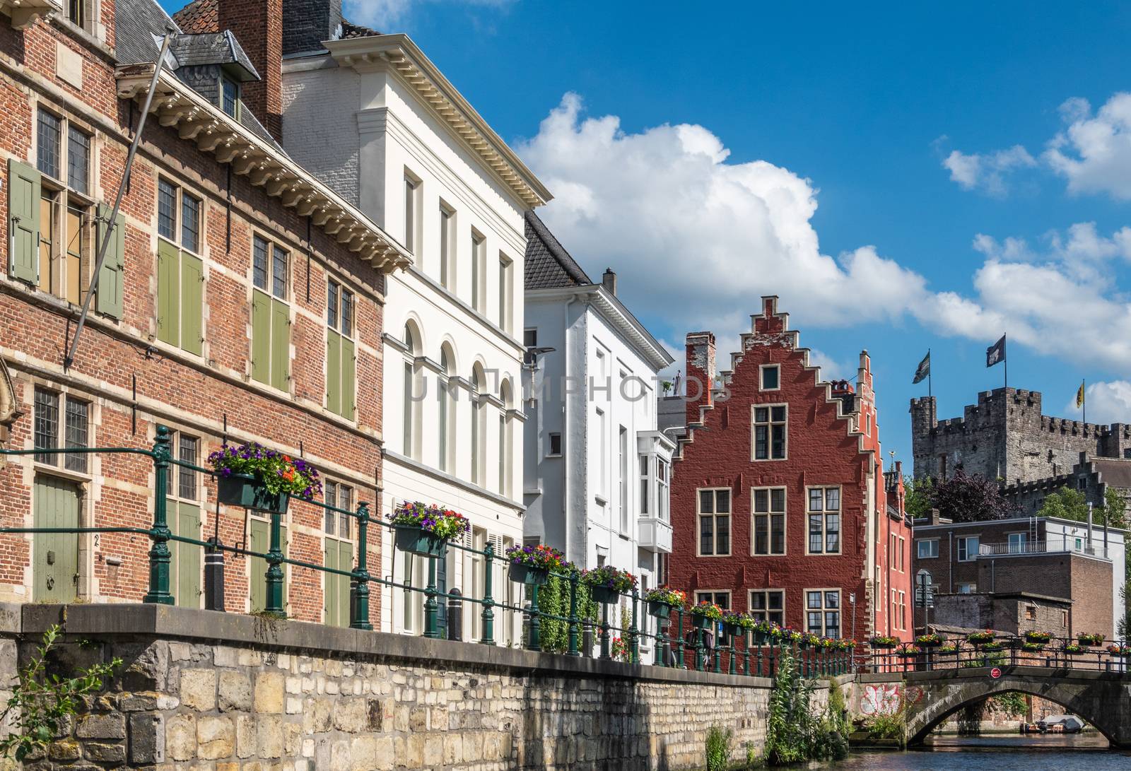 Gent, Flanders, Belgium -  June 21, 2019: Narrow Lieve River leads along old bourgeois mansions to Gravensteen Castle under blue sky with white clouds. Bluish water. Flowers on quays.