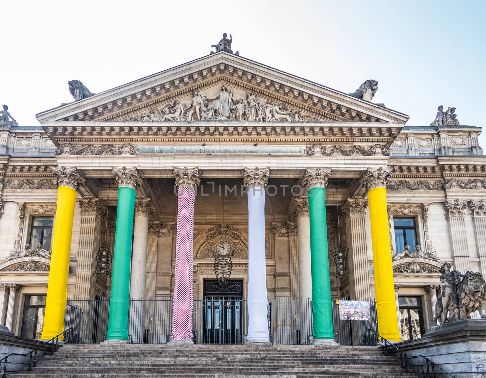 Brussels, Belgium -  June 22, 2019: Brown stone Front facade with tall columns and tympanum with statues on top. Pillars wrapped in bright colors of the Stock Exchange building, Bourse de Bruxelles . Silver sky.