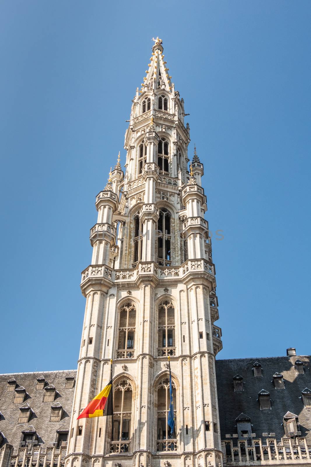 Brussels, Belgium - June 22, 2019: Closeup of gray stone spire of city hall with Belgian flag on Grand Place against blue sky. Golden Saint Michael statue on top.