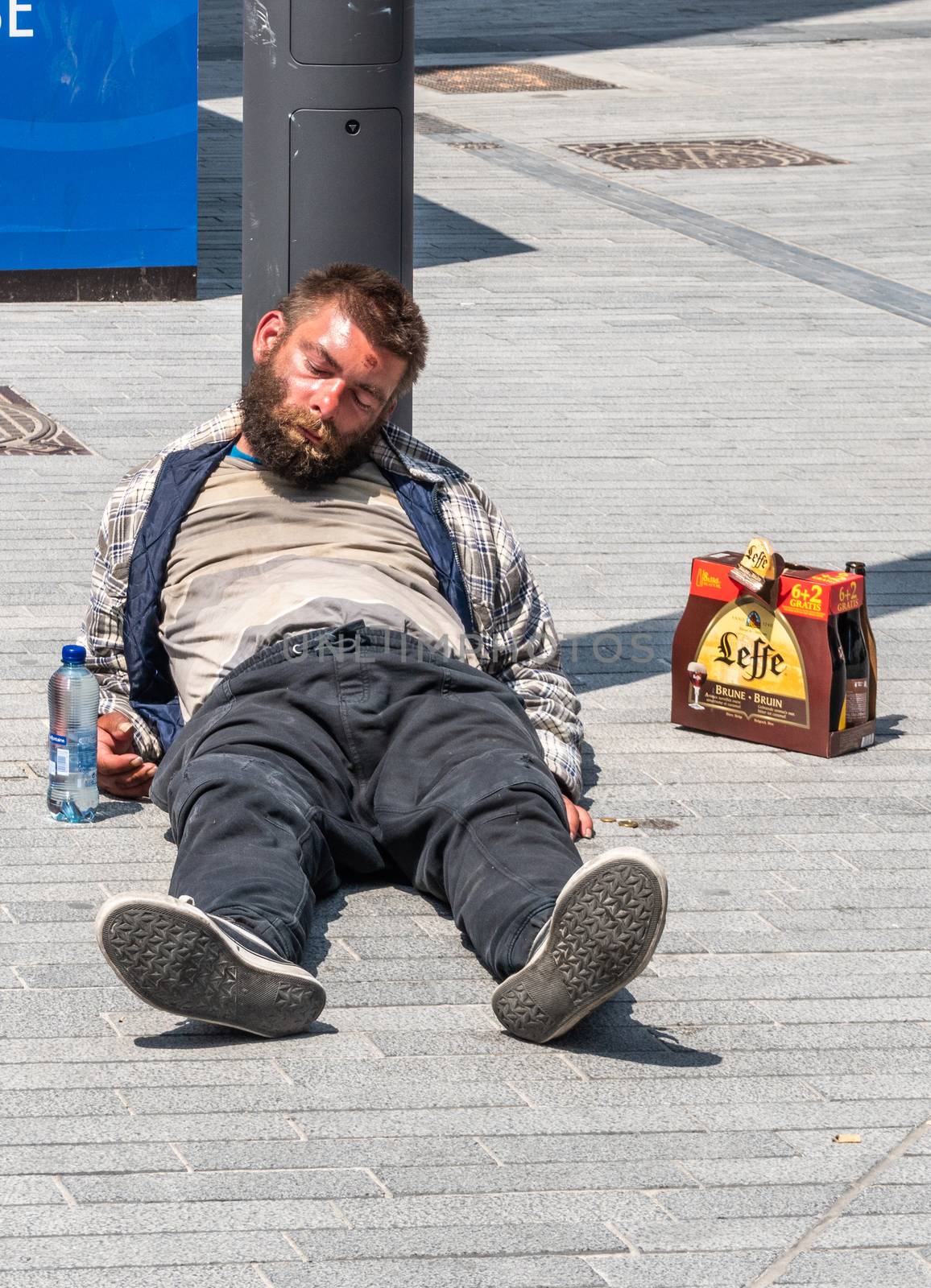 Brussels, Belgium - June 22, 2019: Filthy drunken lad sleeps in sun on gray stone Boulevard Anspach with six-pack of Leffe Abbey ale beers besides him.