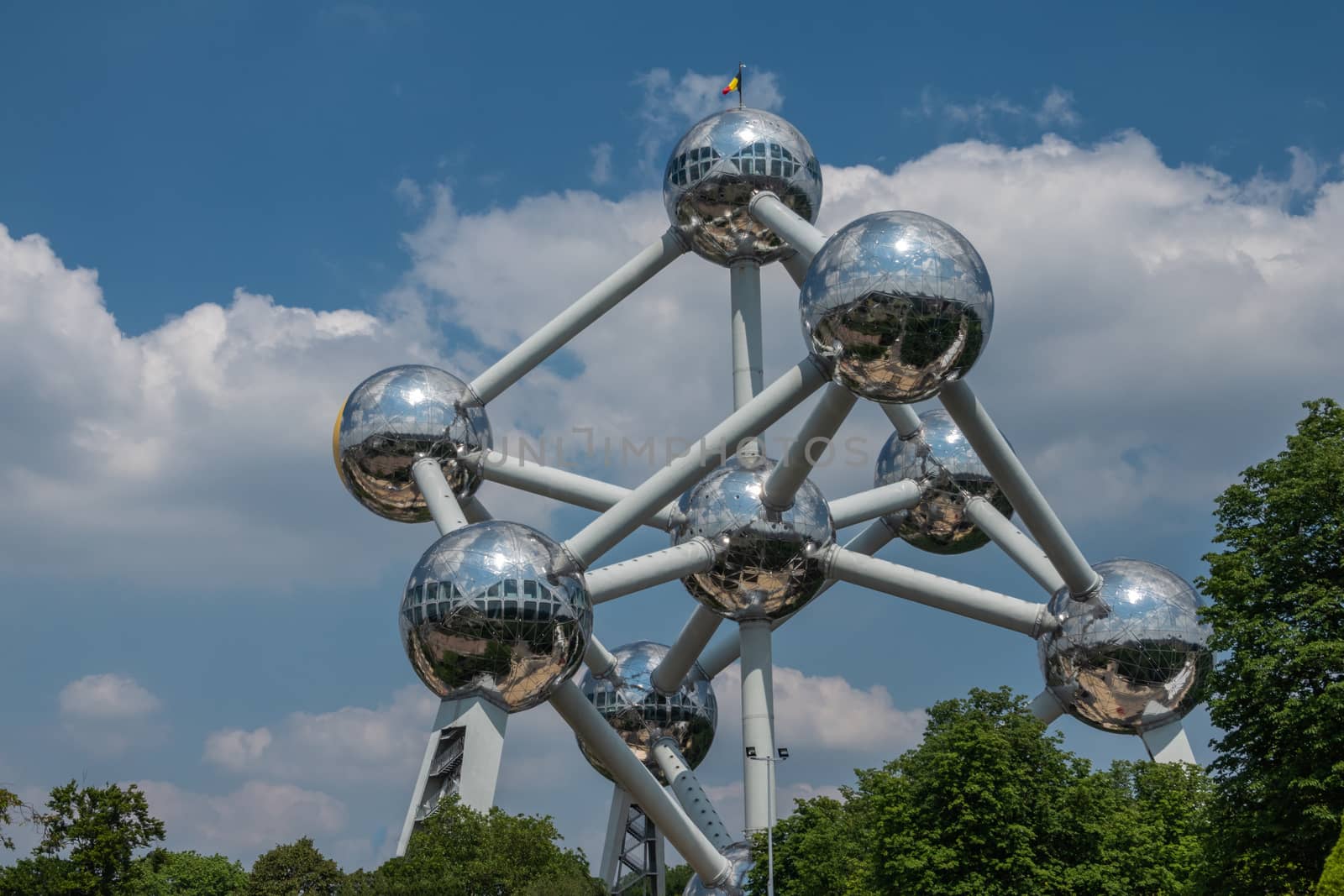 Brussels, Belgium - June 22, 2019: Atomium monument with its silver balls and pipes against blue sky with white cloudscape and green foliage on bottom.