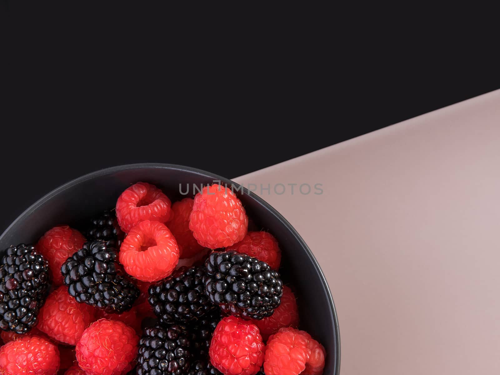 Blackberries and raspberries in a black bowl. Black and light pink background. Top view.