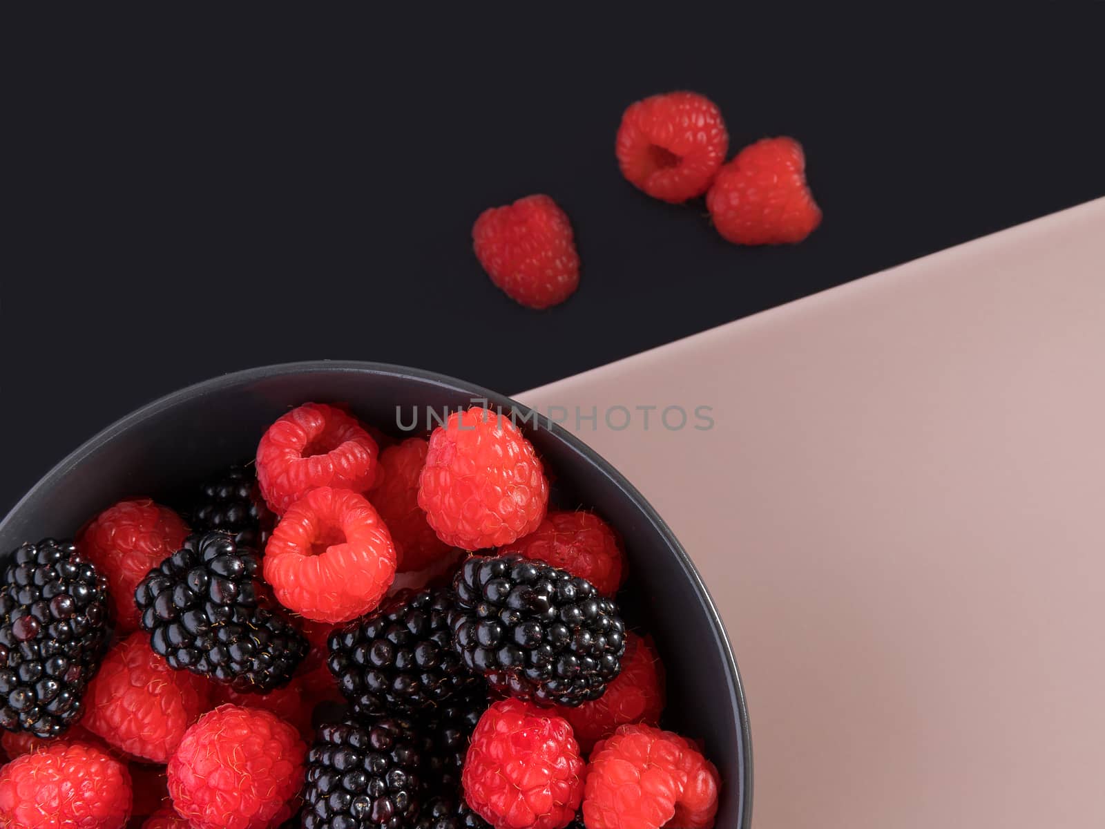 Blackberries and raspberries in a black bowl. Black and light pink background. Top view.