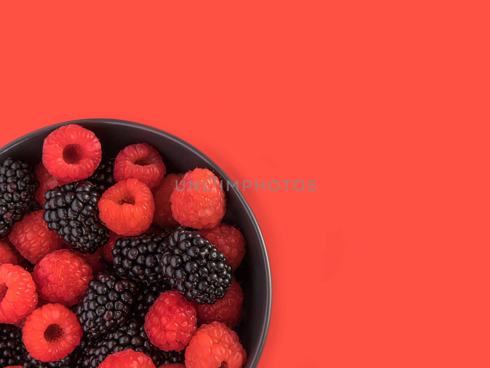 Blackberries and raspberries in a black bowl. Red background. Top view.